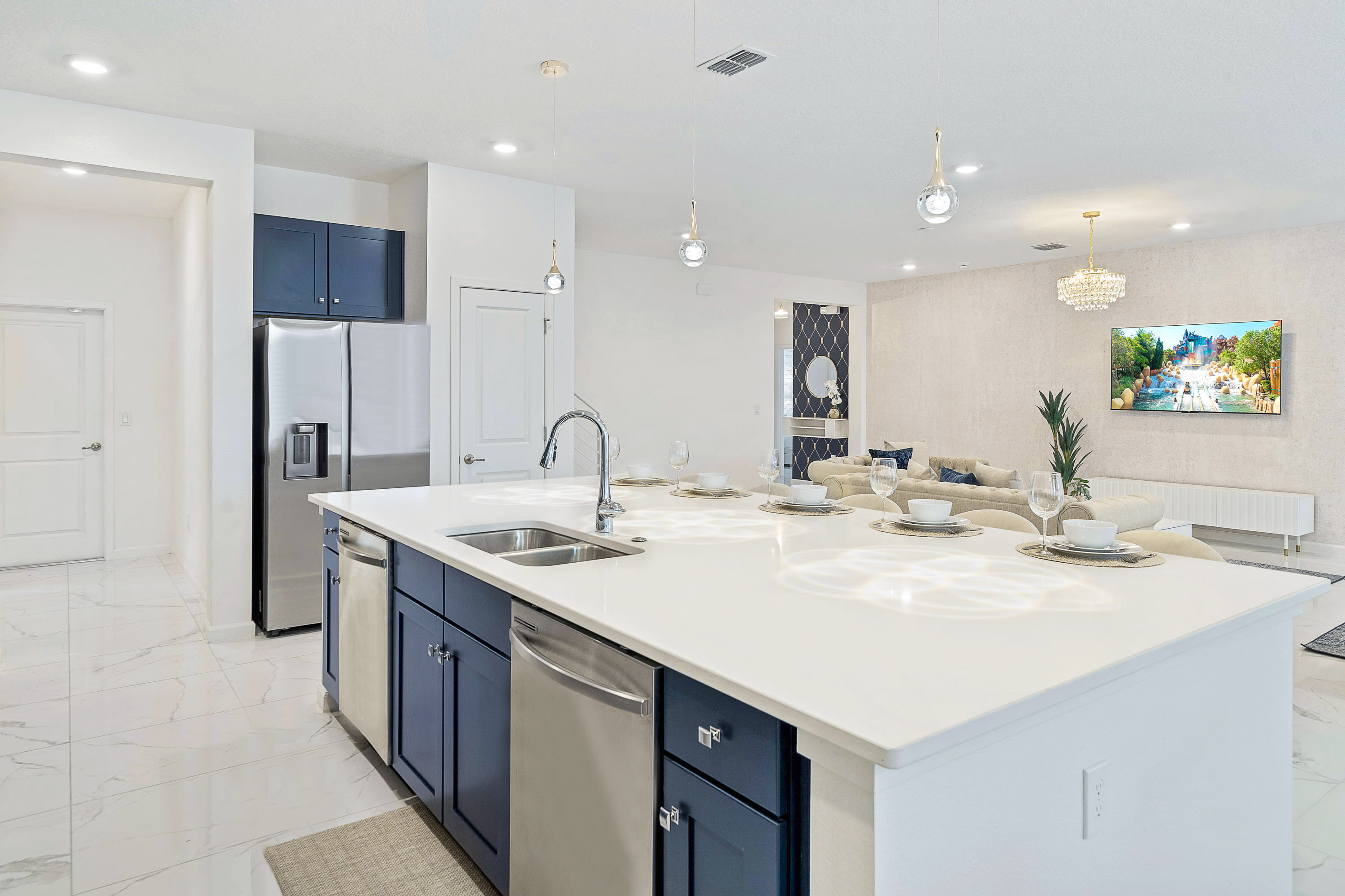 A spacious kitchen island with a double sink and white bar stools, featuring dark blue lower cabinets and a stainless steel dishwasher, with a view into the bright, open-concept living area.