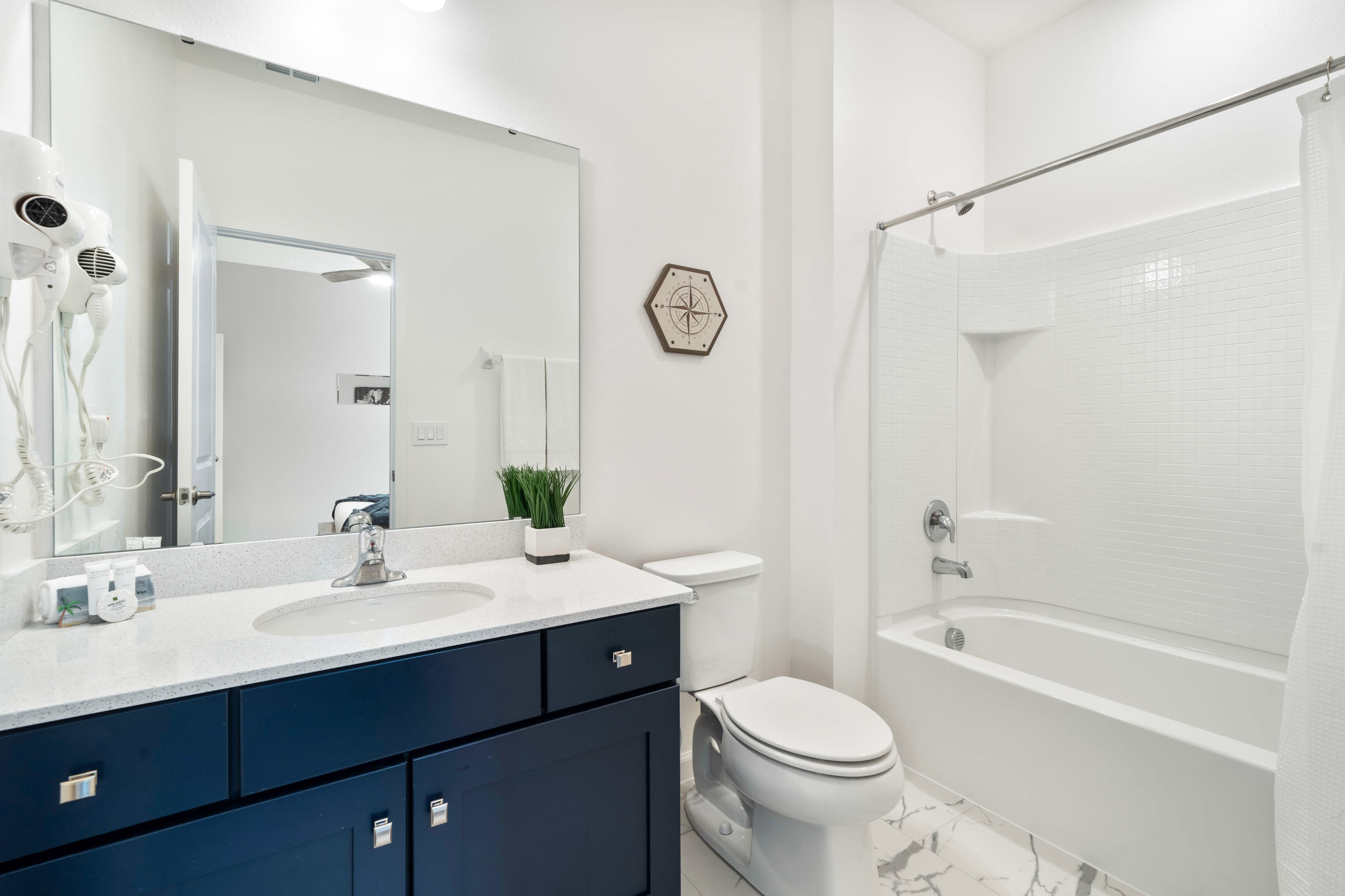 Bright bathroom with a dark blue vanity, white countertop, and single sink, a toilet, and a shower-tub combo with white subway tile. A compass-themed wall decor piece hangs above the toilet, and a mirror reflects a bedroom.