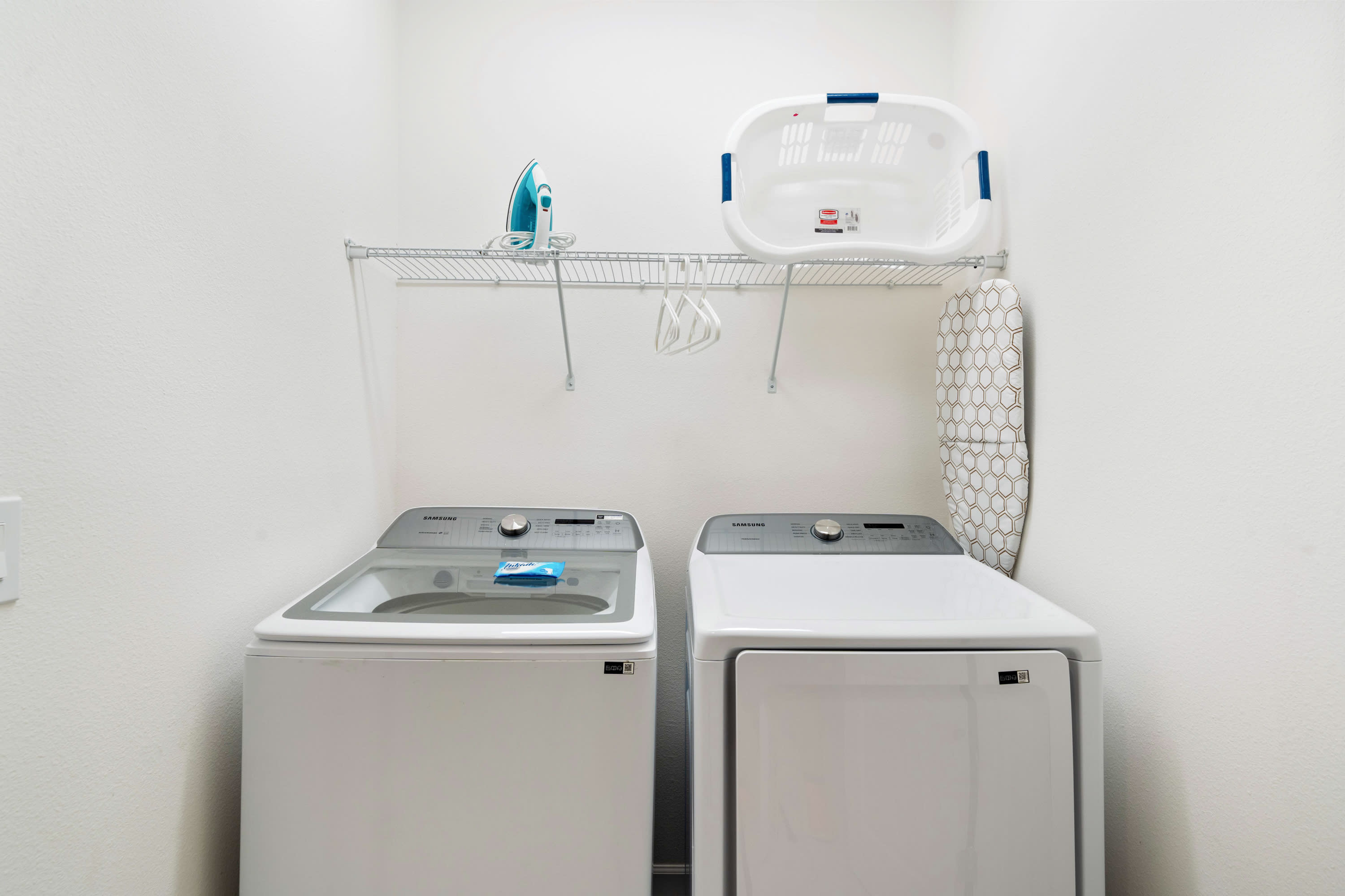 A dedicated laundry area equipped with a top-loading washing machine and a front-loading dryer. A wire shelf above holds a laundry basket, iron, and ironing board, providing essential amenities for guests.