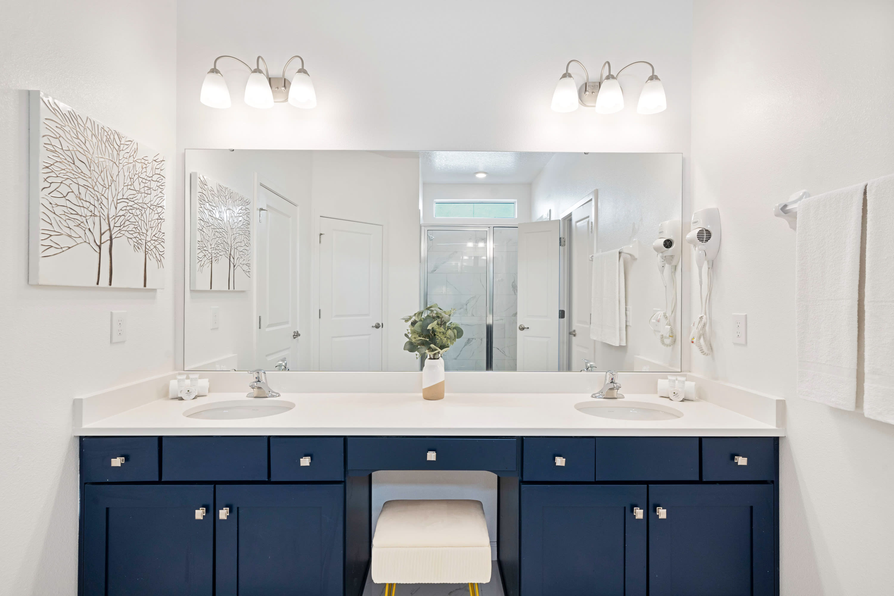 A spacious white bathroom with a long double vanity featuring dark blue cabinets, white quartz countertops, and a large mirror. A white stool is tucked under the vanity, and a plant decorates the counter.