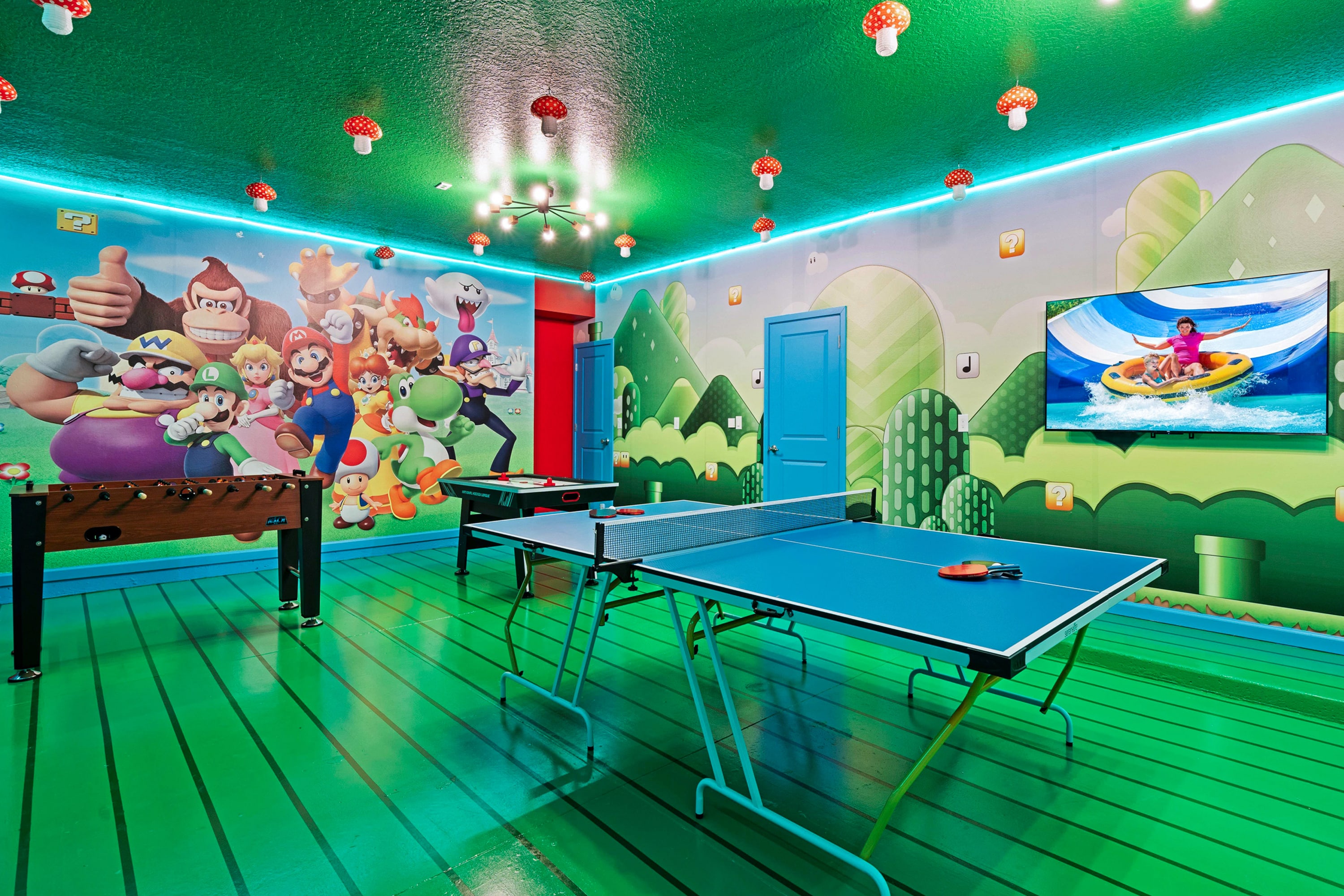 Vibrant game room with a Super Mario Bros. theme, featuring a foosball table, ping pong table, and a wall-mounted TV, all under a green ceiling with mushroom lights.