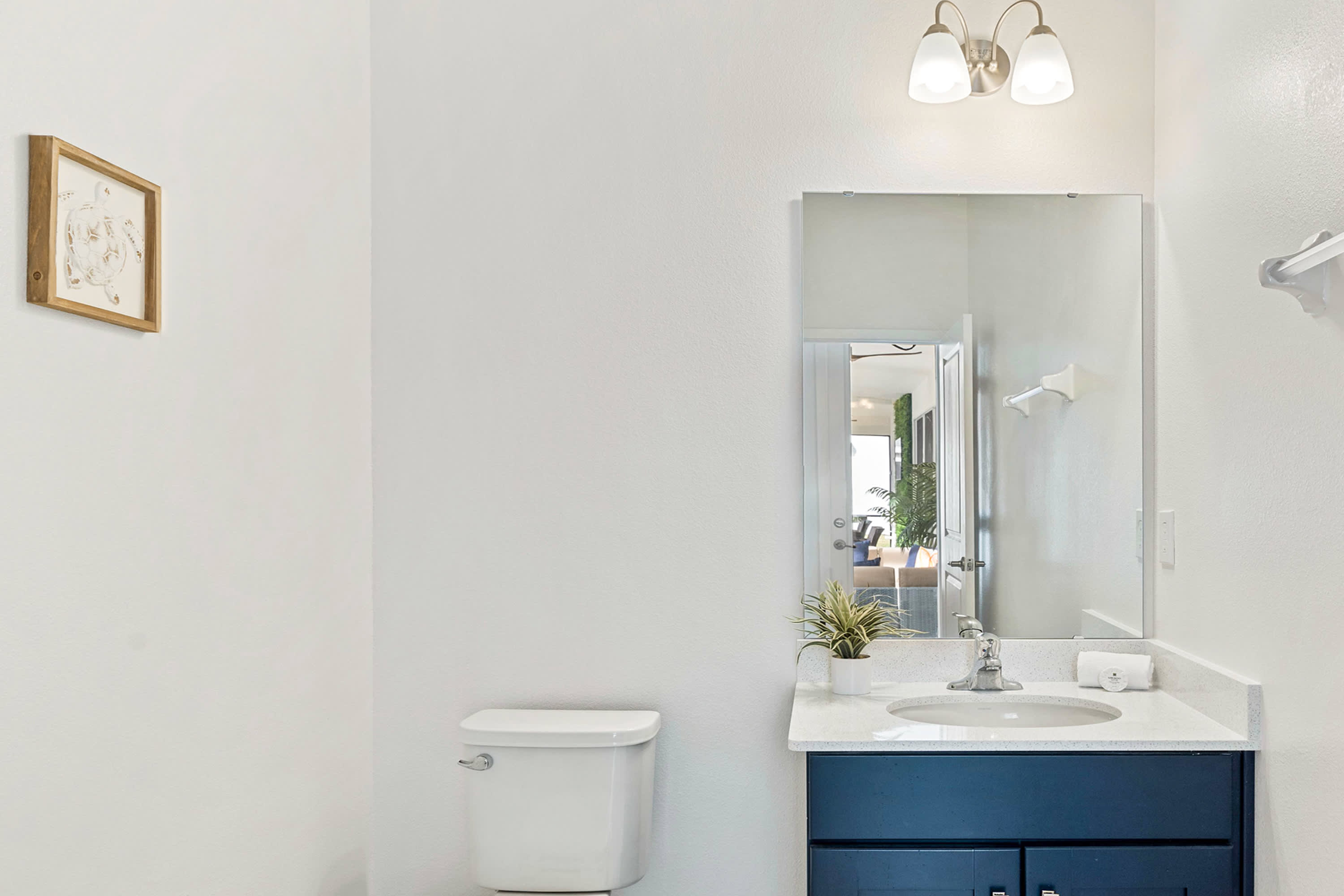 A compact and bright bathroom featuring a white toilet and a single vanity with a dark blue cabinet and white countertop. A small framed artwork hangs on the wall, and a mirror reflects the simple, clean design.