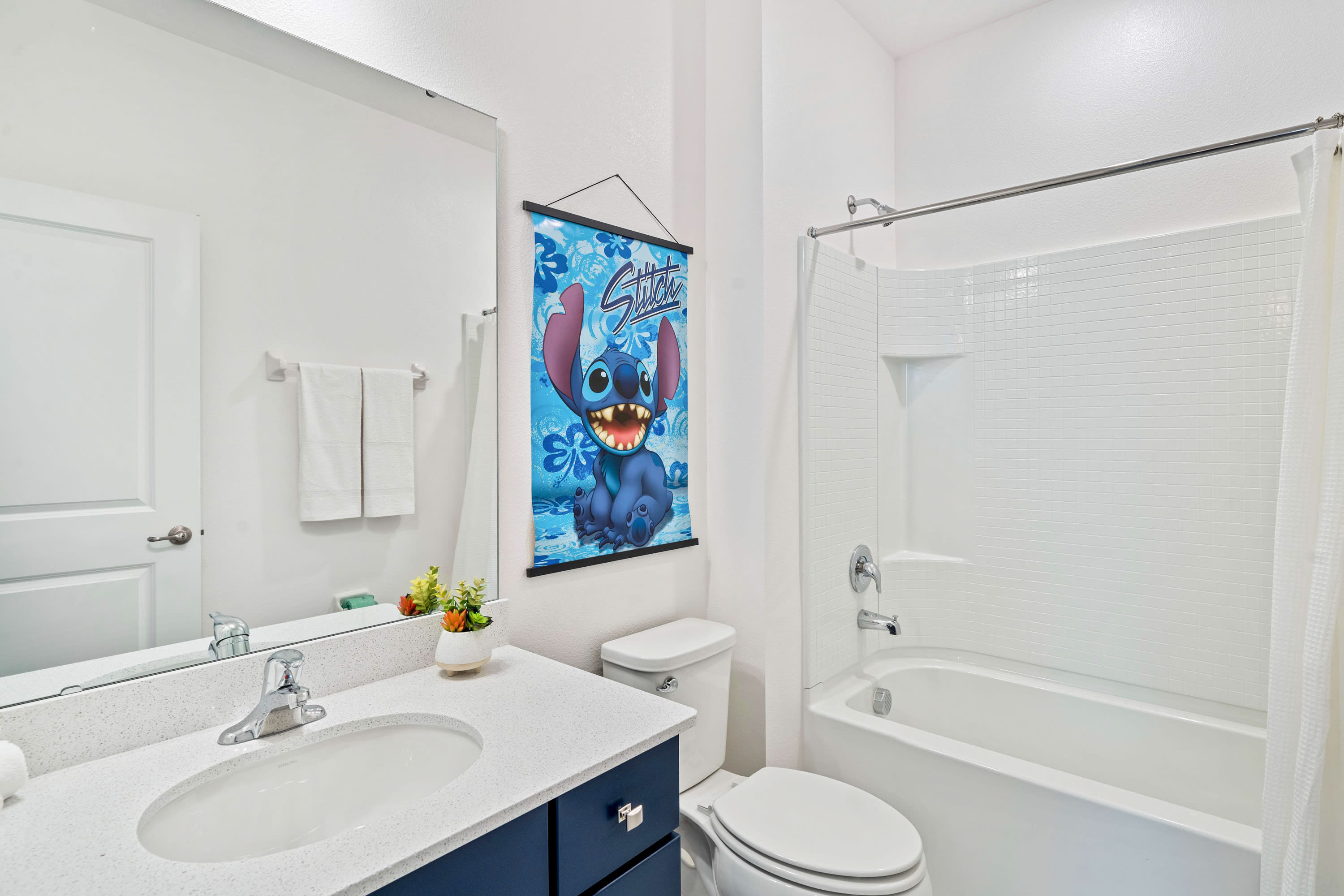 Clean white bathroom featuring a tub/shower combo with subway tile surround and a single vanity with a dark blue cabinet and white countertop. A large Stitch banner hangs on the wall above the toilet, reinforcing the room's theme.
