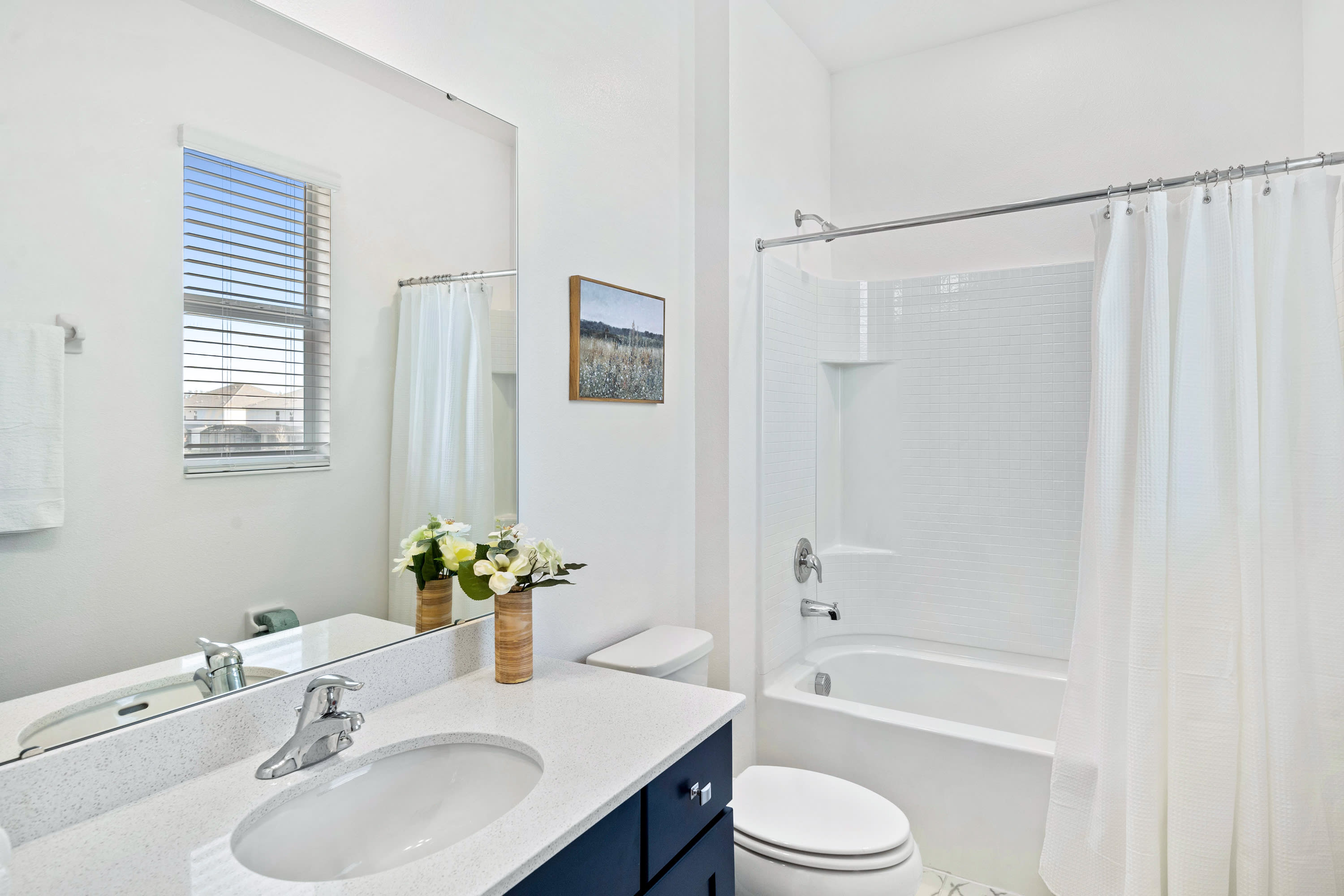 A bright white bathroom with a single dark blue vanity, white countertop, and a large mirror. It features a toilet and a bathtub with a shower, and a window above the vanity.