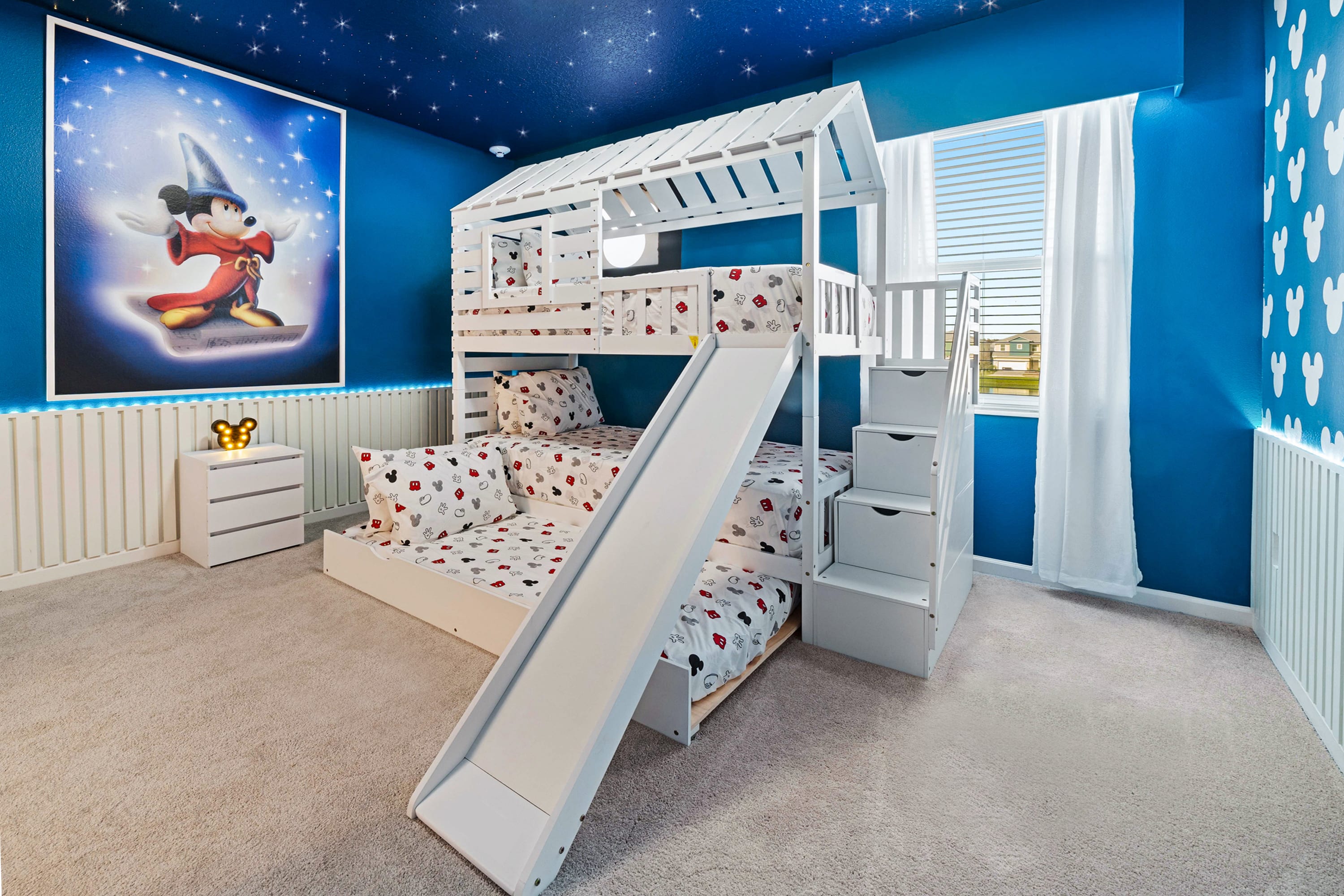 A playful Mickey Mouse themed bedroom featuring a white house-shaped bunk bed with a slide and stairs. The room has bright blue walls, a starry night ceiling, and a large framed Mickey Mouse artwork, creating a magical atmosphere.