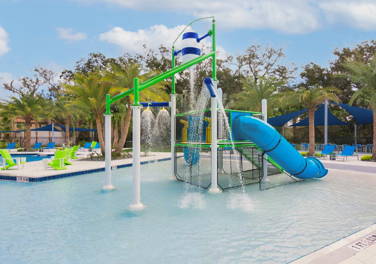 A close-up view of the colorful splash pad area, highlighting a blue water slide, a large tipping bucket, and multiple water jets. Bright green lounge chairs and palm trees are visible around the edges of the shallow water play zone.
