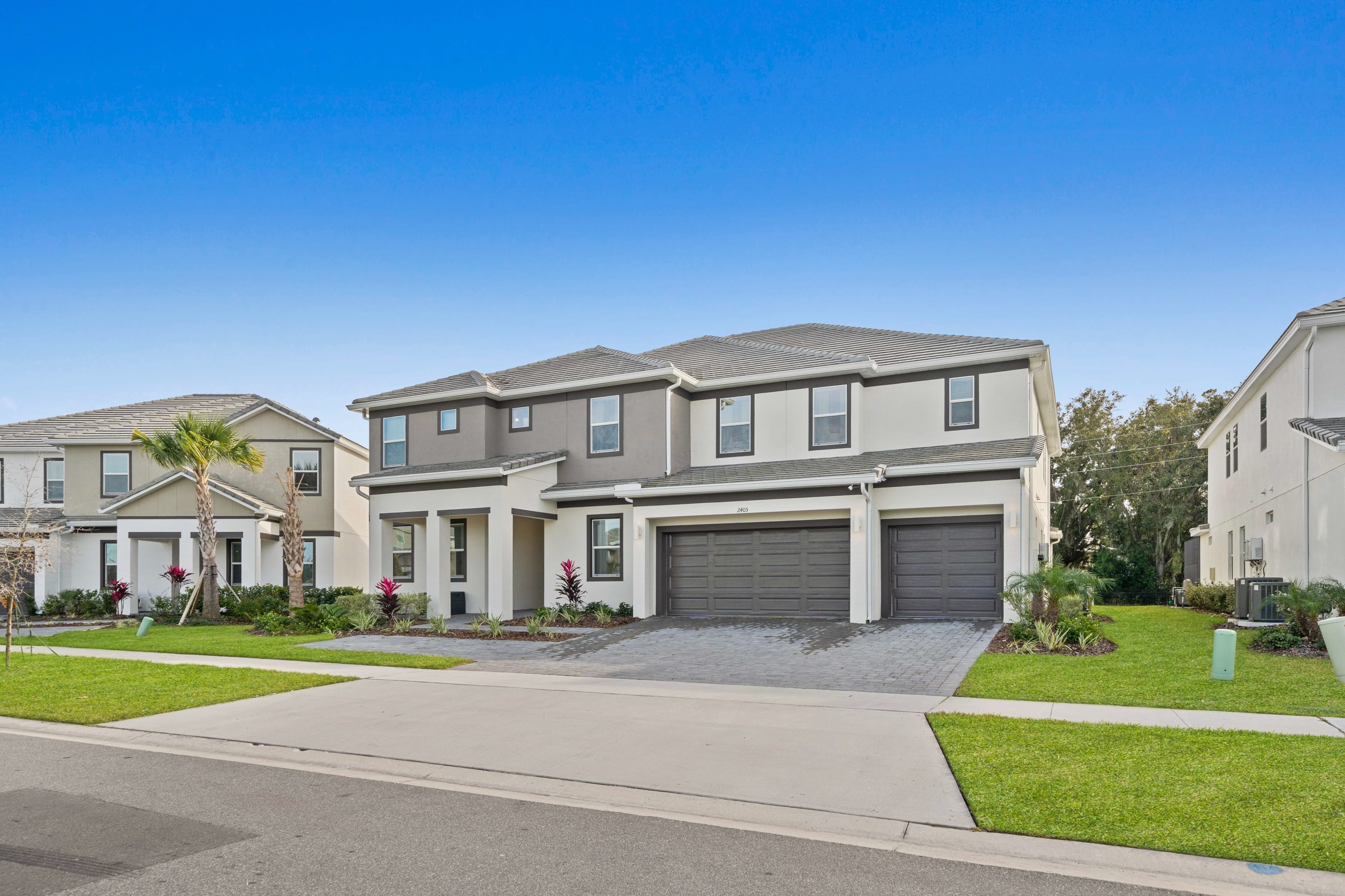 The front exterior of a large, modern two-story house with a light gray and white facade, dark gray garage doors, and a paved driveway. Lush green lawns and palm trees frame the property under a clear blue sky.