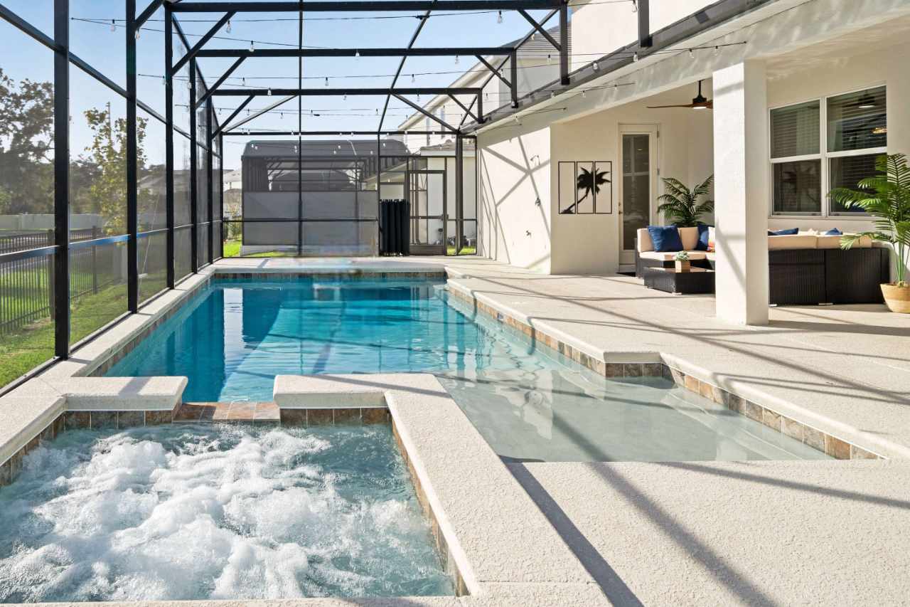 Screened outdoor pool area featuring a rectangular swimming pool and an adjacent bubbling hot tub, surrounded by a light-colored concrete deck and an outdoor seating area.