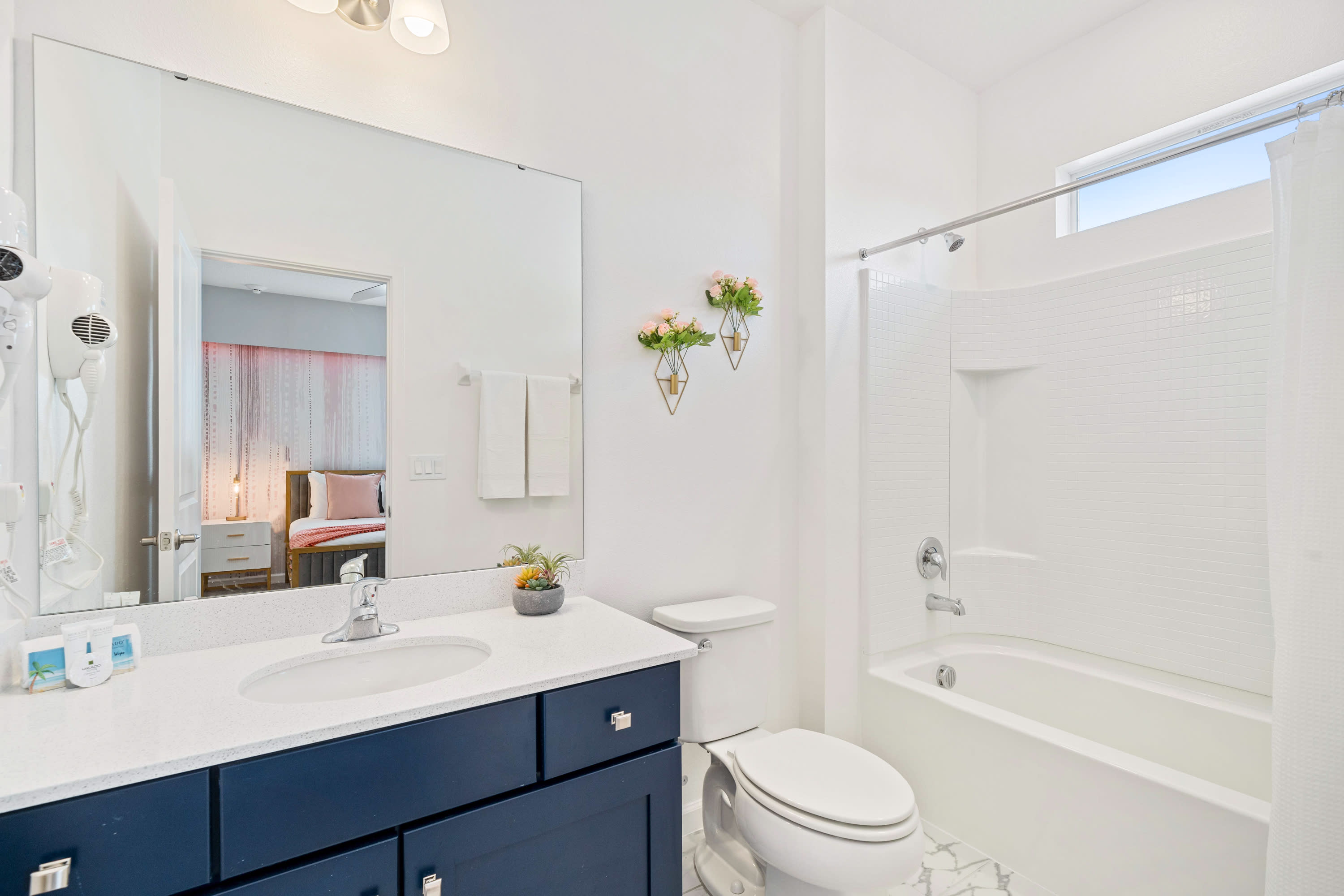 Bright white bathroom featuring a dark blue vanity with a white countertop and single sink, a toilet, and a shower-tub combo with white subway tile. A large mirror reflects a bedroom with a bed and pink wall art.