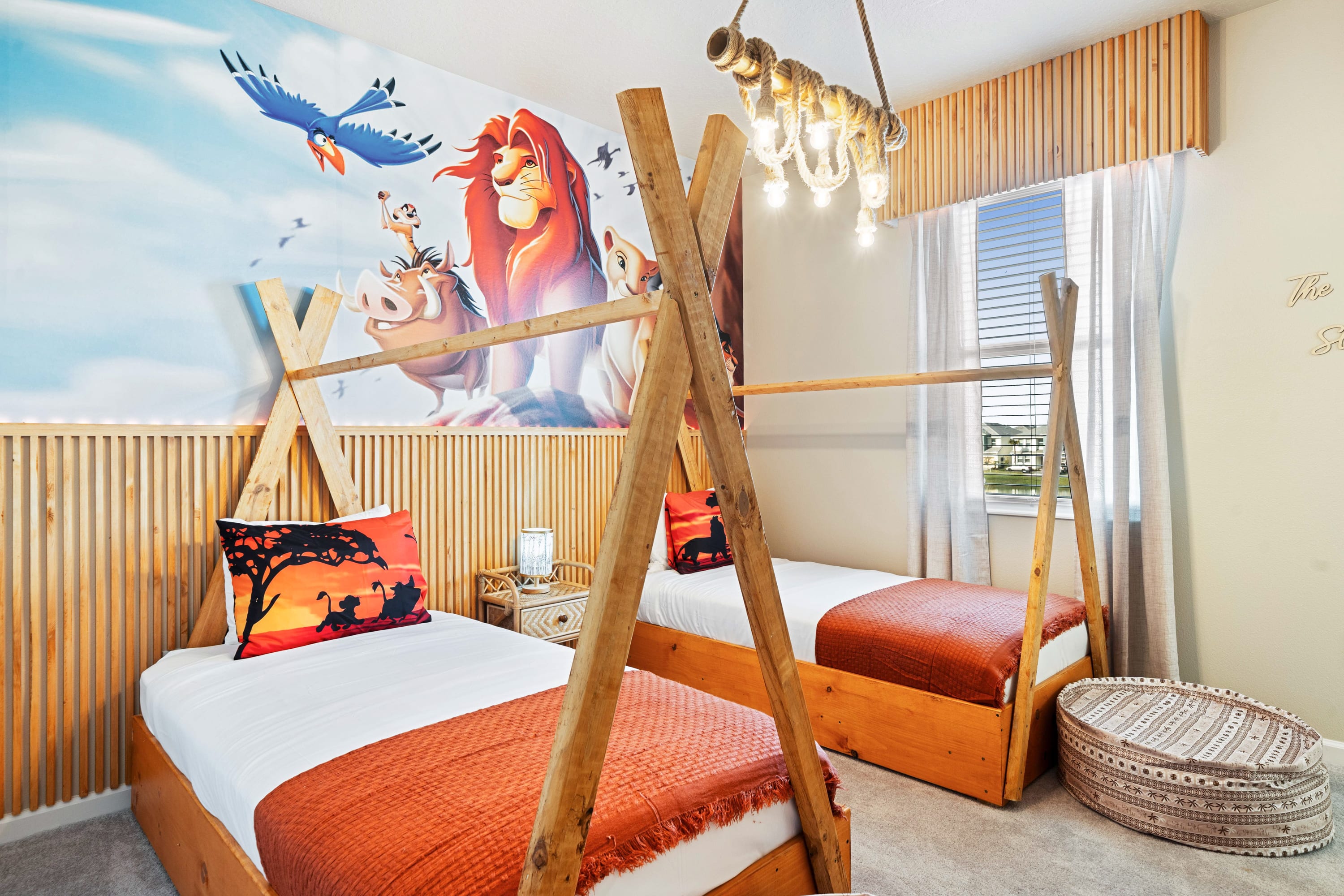 Vibrant kids' bedroom featuring two twin beds with unique wooden teepee-style frames and orange bedding, set against a colorful Lion King mural. A rustic chandelier hangs from the ceiling, adding to the playful theme.