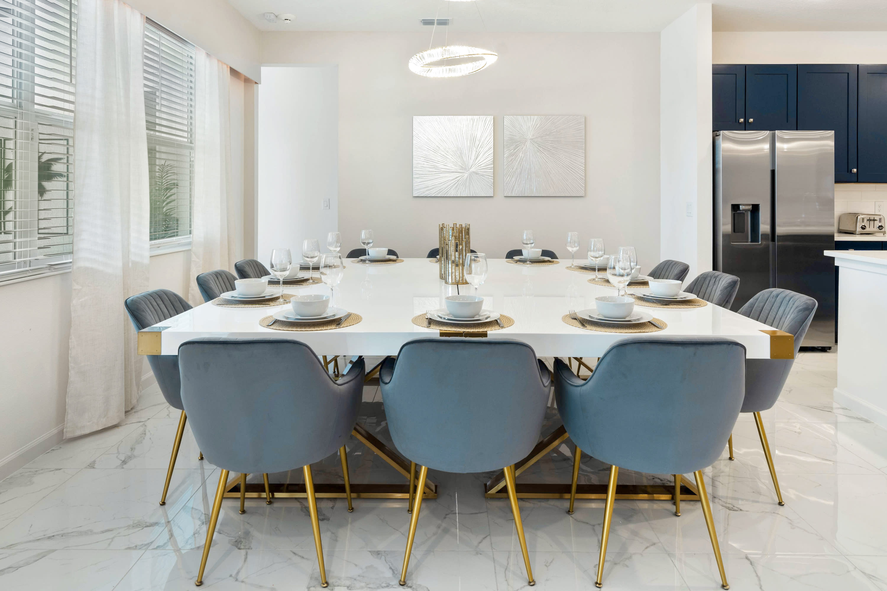 A modern dining area centered around a large white rectangular table with gold accents, surrounded by grey velvet chairs with gold legs, set beneath a contemporary chandelier and flanked by abstract silver wall art.