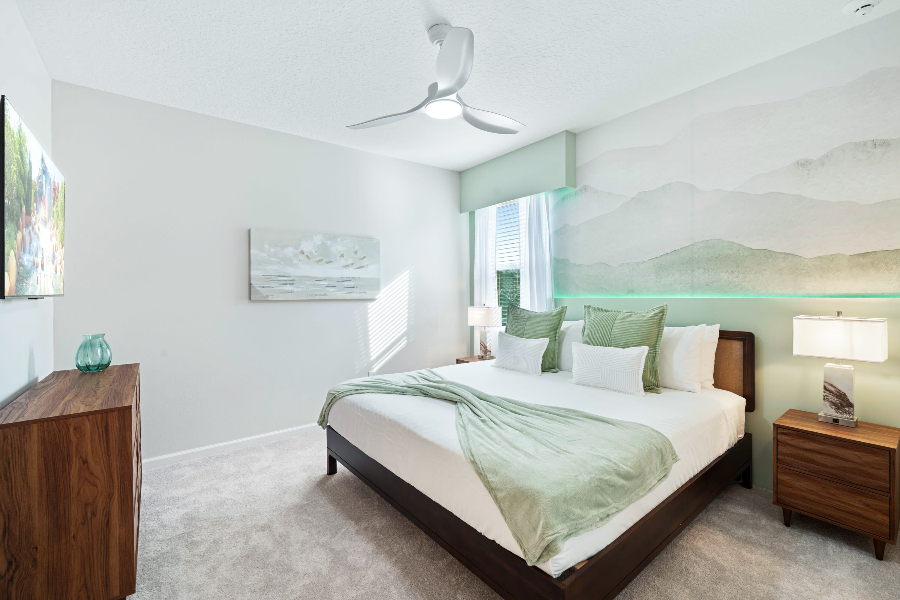Serene bedroom with a king-sized bed featuring a dark wood frame, light green bedding, and a decorative mountain mural with ambient lighting above the headboard. A ceiling fan and a wooden dresser with a TV are also visible.