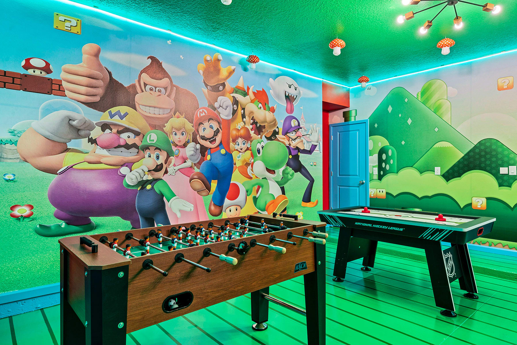 Themed game room with Super Mario Bros. wall art and a green floor, featuring a wooden foosball table and a black air hockey table, illuminated by mushroom-shaped ceiling lights.