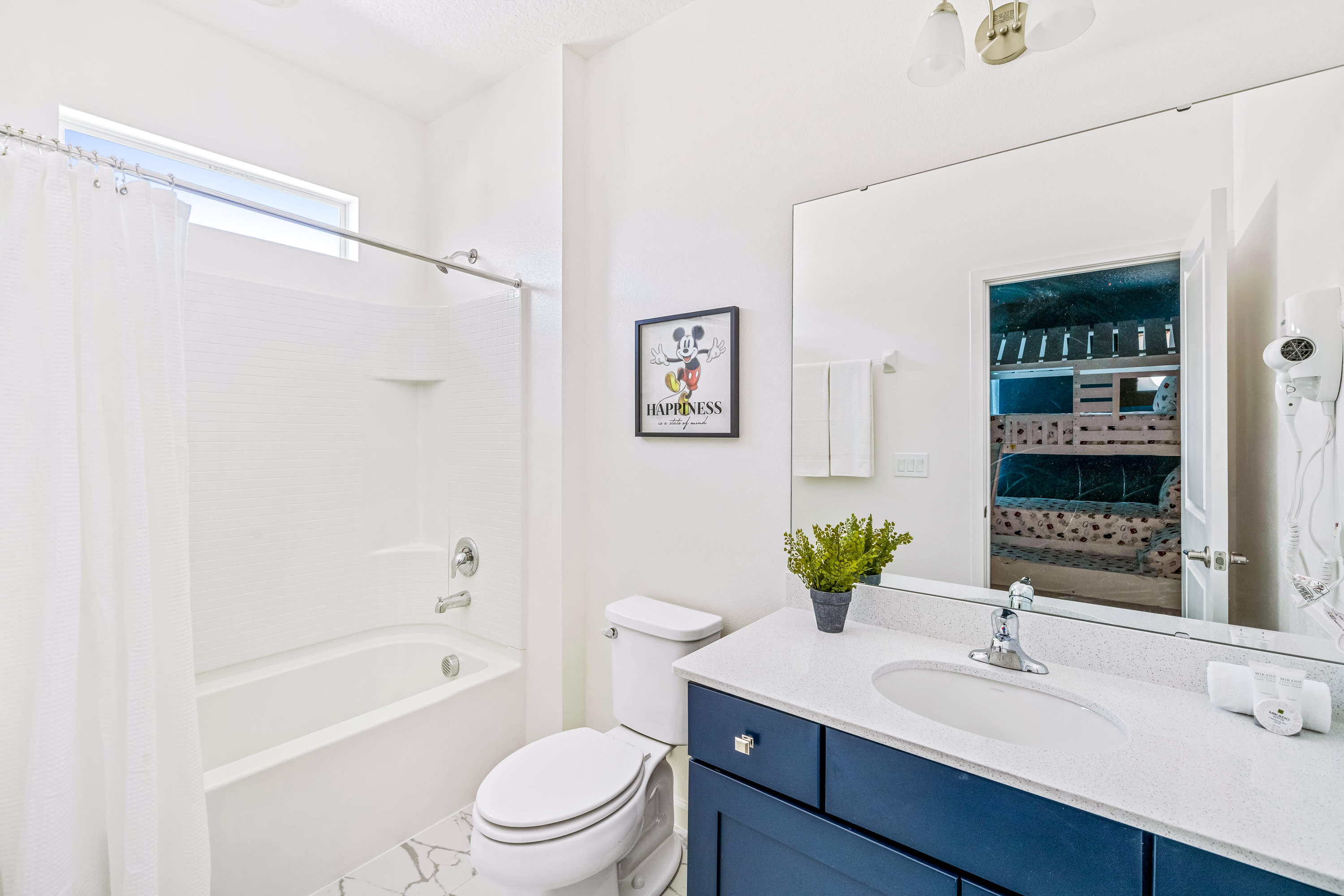 Bright and modern bathroom with a white tub/shower combo and a single vanity featuring a dark blue cabinet and white countertop. A framed Mickey Mouse 'HAPPINESS' art piece adds a cheerful touch, and a large mirror reflects the bunk bed room.