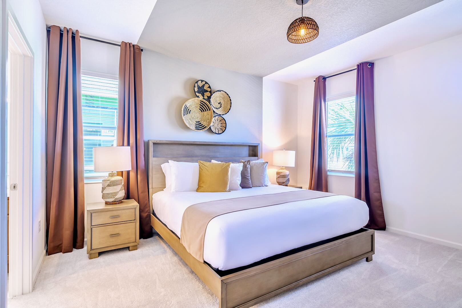 Spacious bedroom featuring a king-sized bed with a light wood headboard, two matching nightstands with lamps, and rich brown curtains. The room has a tray ceiling with a woven pendant light fixture.
