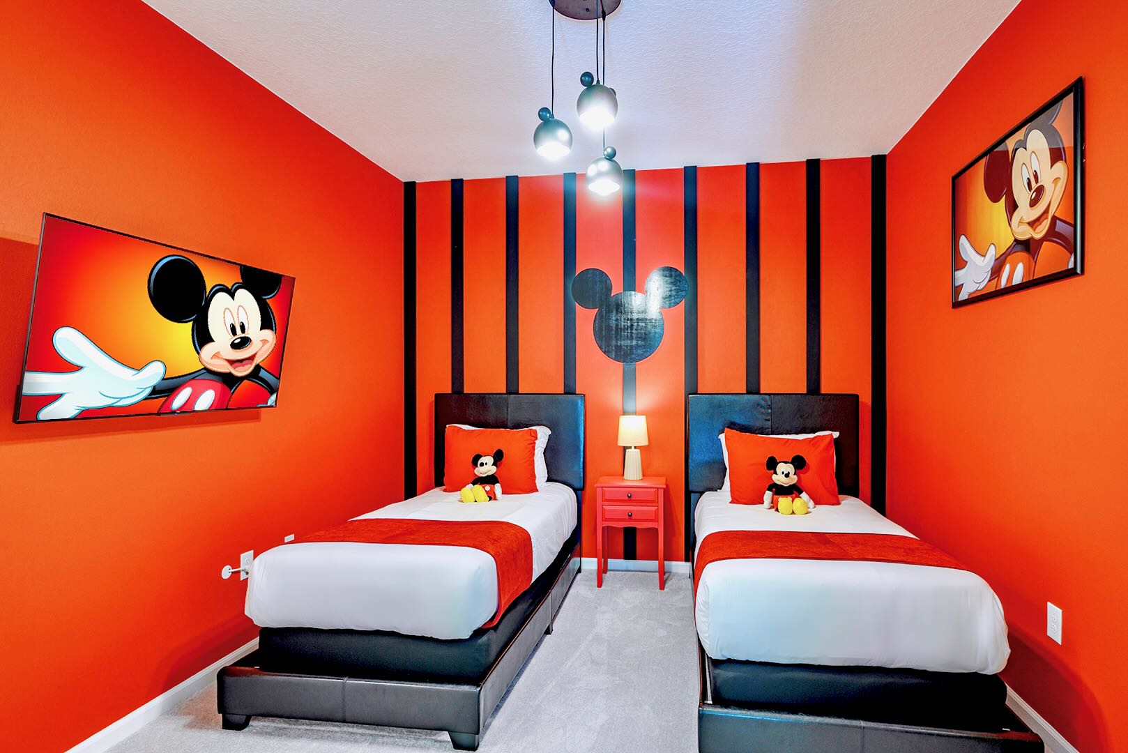 A vibrant Mickey Mouse themed bedroom featuring two twin beds with black headboards, red bedding, and Mickey plush toys. The walls are bright orange with bold black vertical stripes and a large Mickey silhouette.