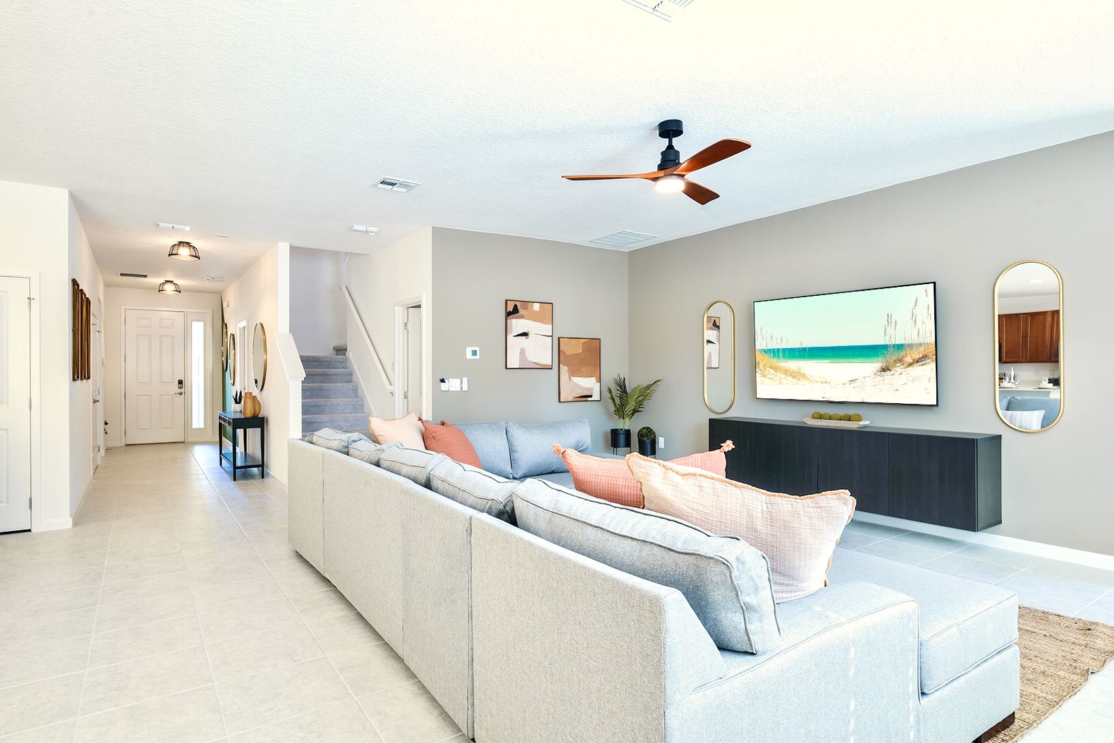 Spacious and bright living room with light grey tile flooring, a large light grey sectional sofa, a wall-mounted TV, and a view towards the front entrance and a carpeted staircase.