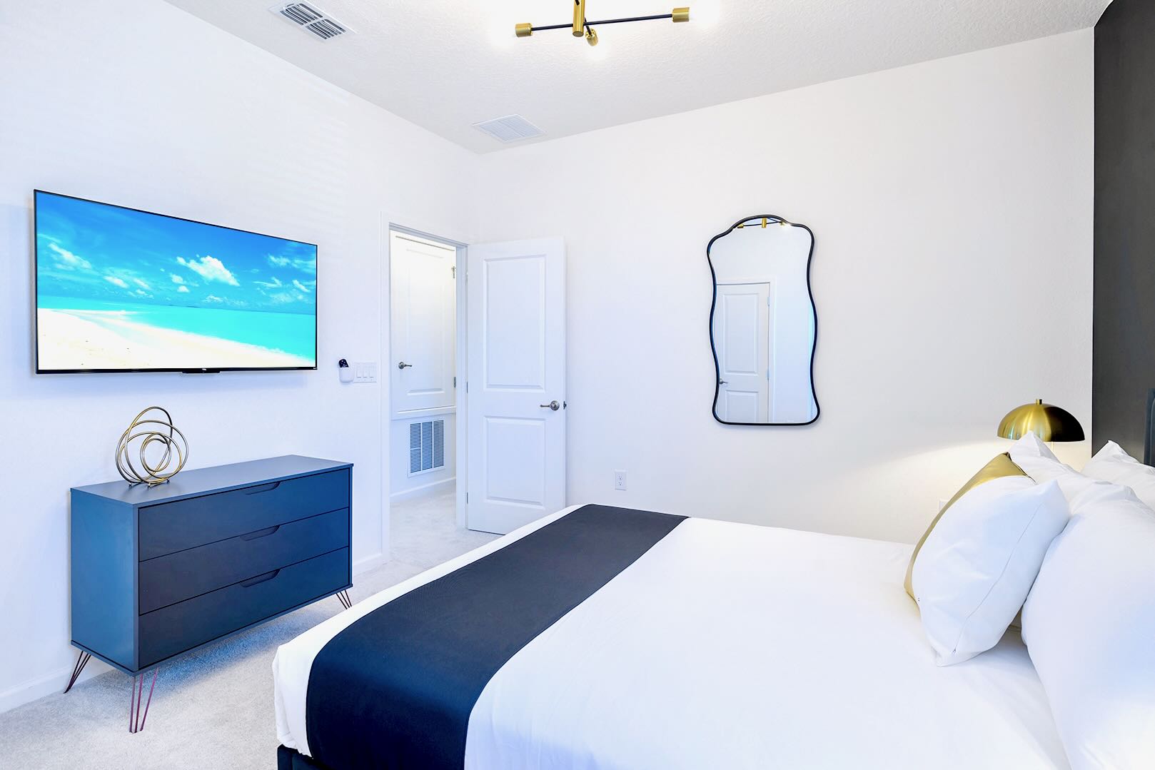 View of the bedroom showing a wall-mounted TV above a dark blue dresser with gold legs, and a decorative mirror on the adjacent wall. A white door leads to another room, all set on light carpet.