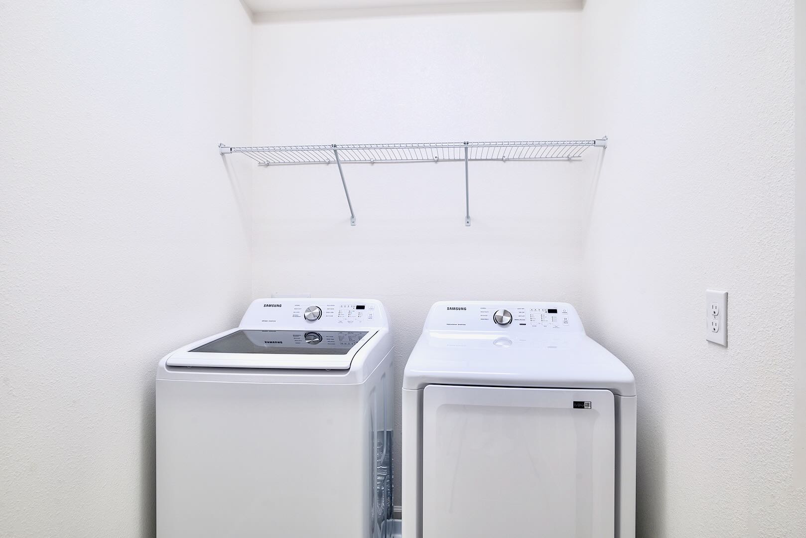 A functional laundry room with clean white walls, featuring a white top-load washing machine and a matching front-load dryer. A wire shelf is mounted above the appliances for convenient storage.