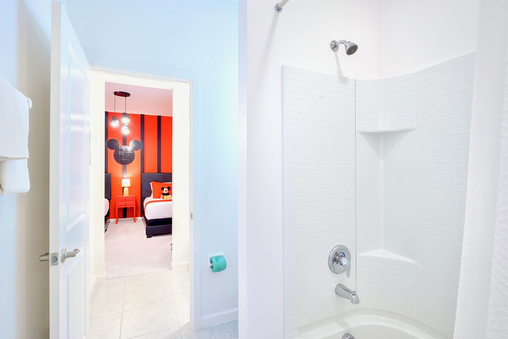 A clean white bathroom featuring a shower/tub combination with white tiled walls and a chrome showerhead. A doorway provides a glimpse into a vibrant orange and black themed bedroom.