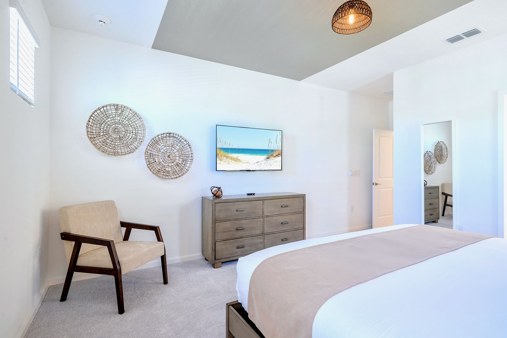 Another view of the bedroom showcasing a wall-mounted TV above a wooden dresser, a comfortable beige armchair, and decorative woven wall art. The room features light carpet and white walls.