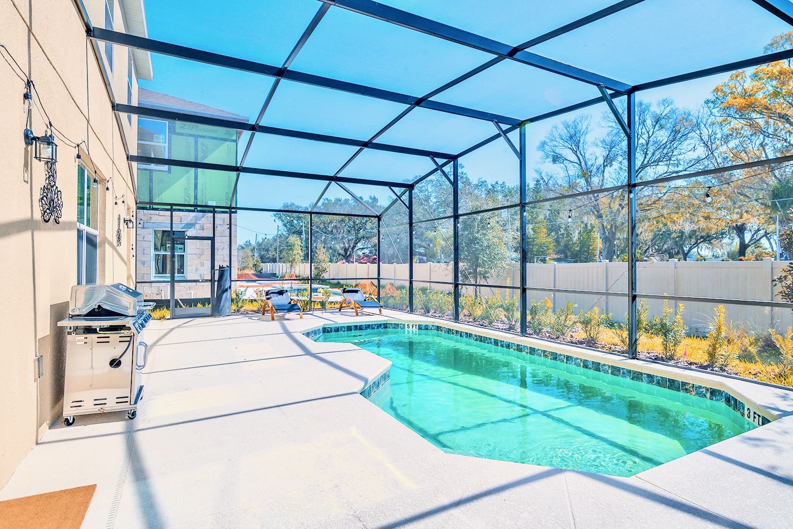 Screened-in private swimming pool with light blue water, surrounded by a concrete deck with lounge chairs and a stainless steel BBQ grill, offering views of a fenced backyard and trees.
