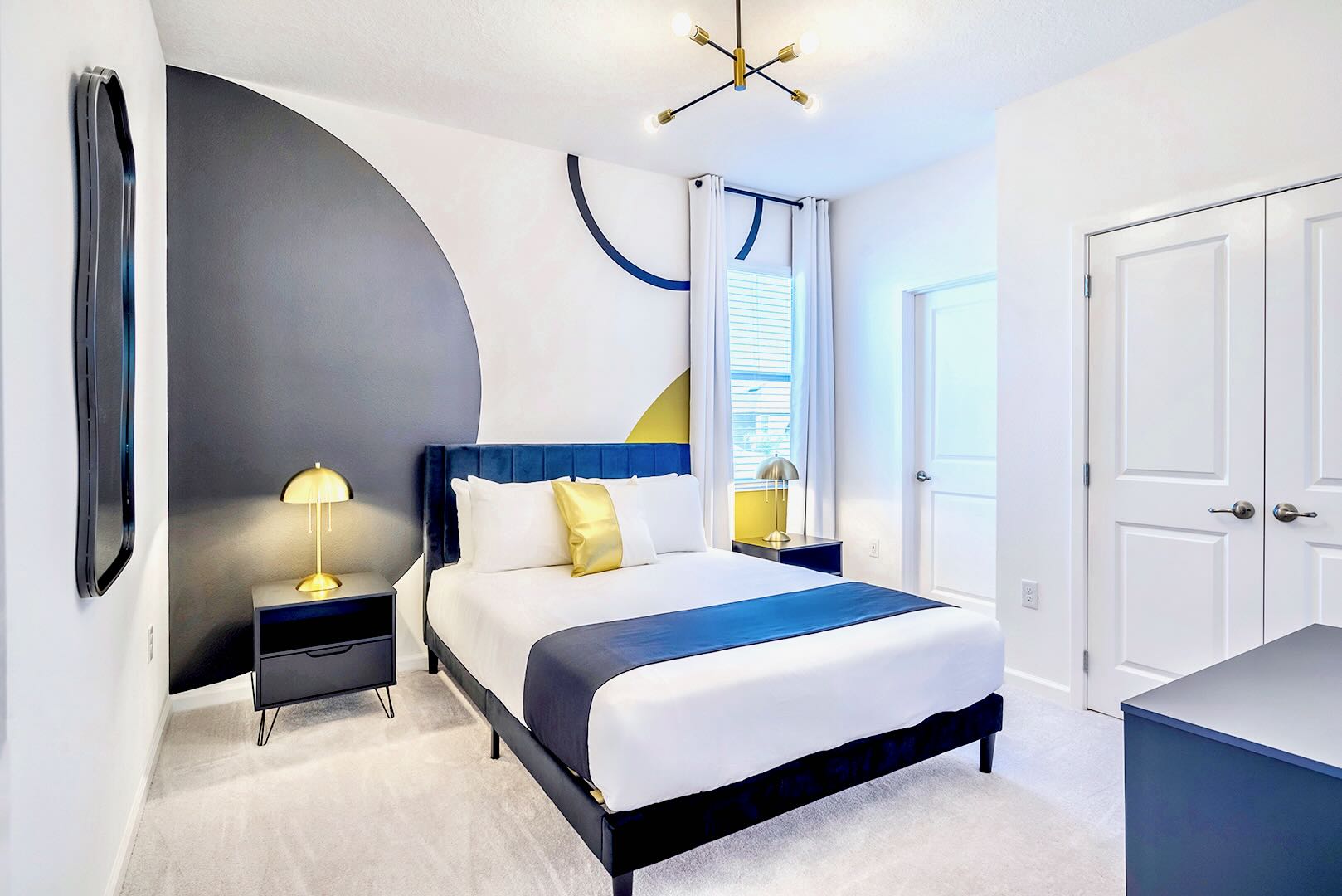Modern bedroom featuring a queen-sized bed with a dark blue velvet headboard and a striking geometric accent wall in black, white, and yellow. Two black nightstands with gold lamps flank the bed.