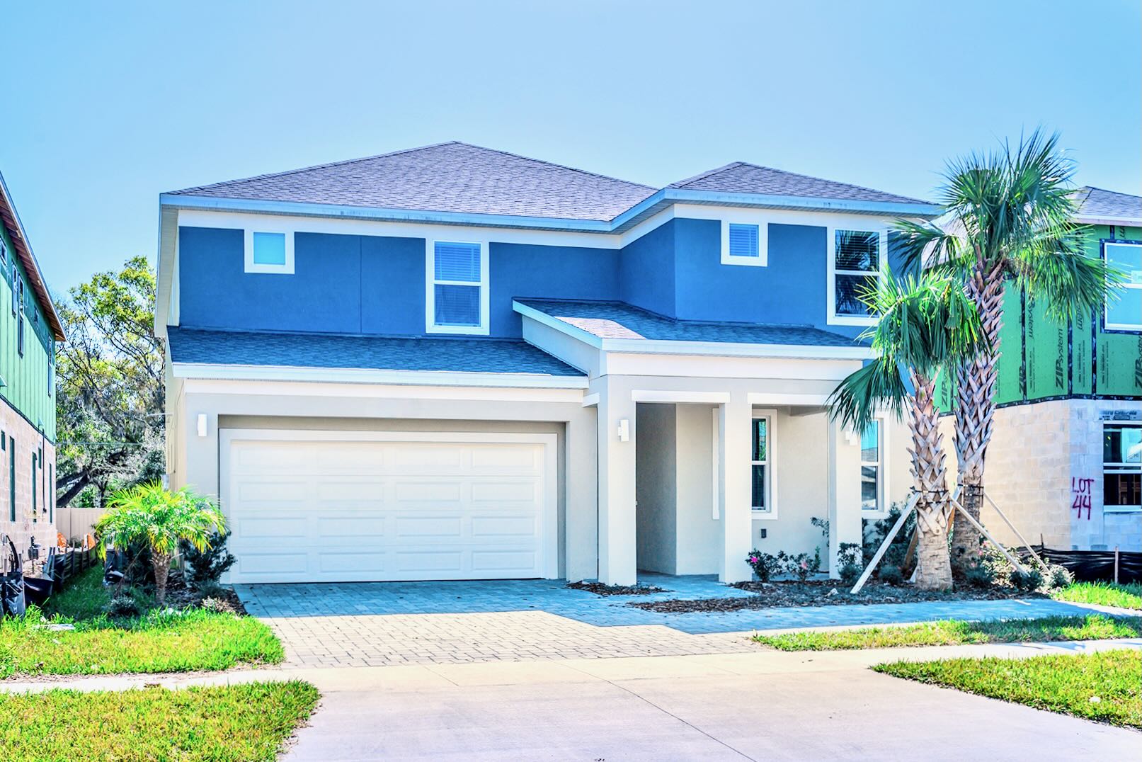 Modern two-story house with a blue and light beige exterior, a two-car garage, a paved driveway, and a small palm tree in the front yard under a clear blue sky.