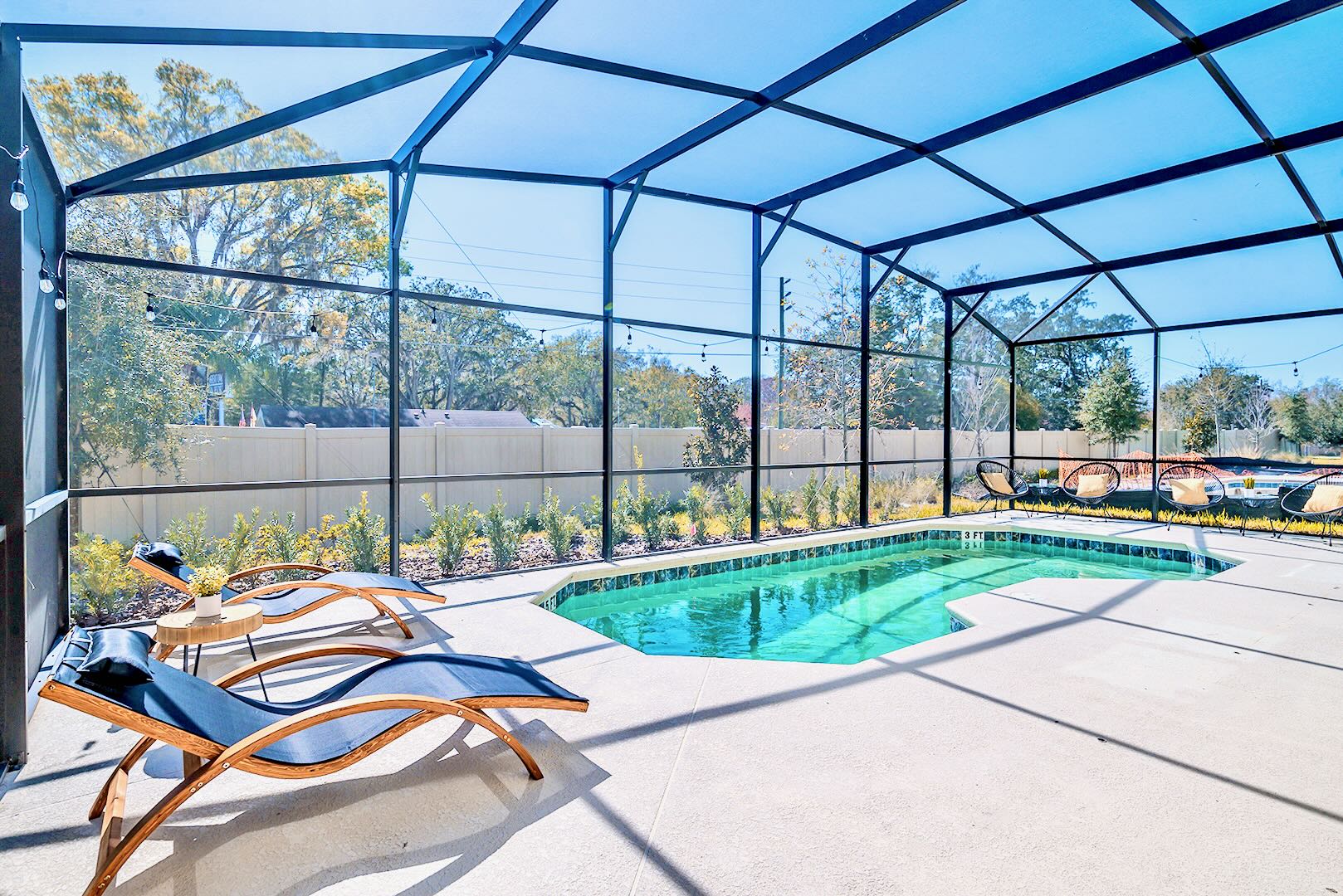 Screened-in private swimming pool with light blue water, surrounded by a concrete deck with two modern wooden lounge chairs and string lights overhead, set against a backdrop of a low fence and trees.