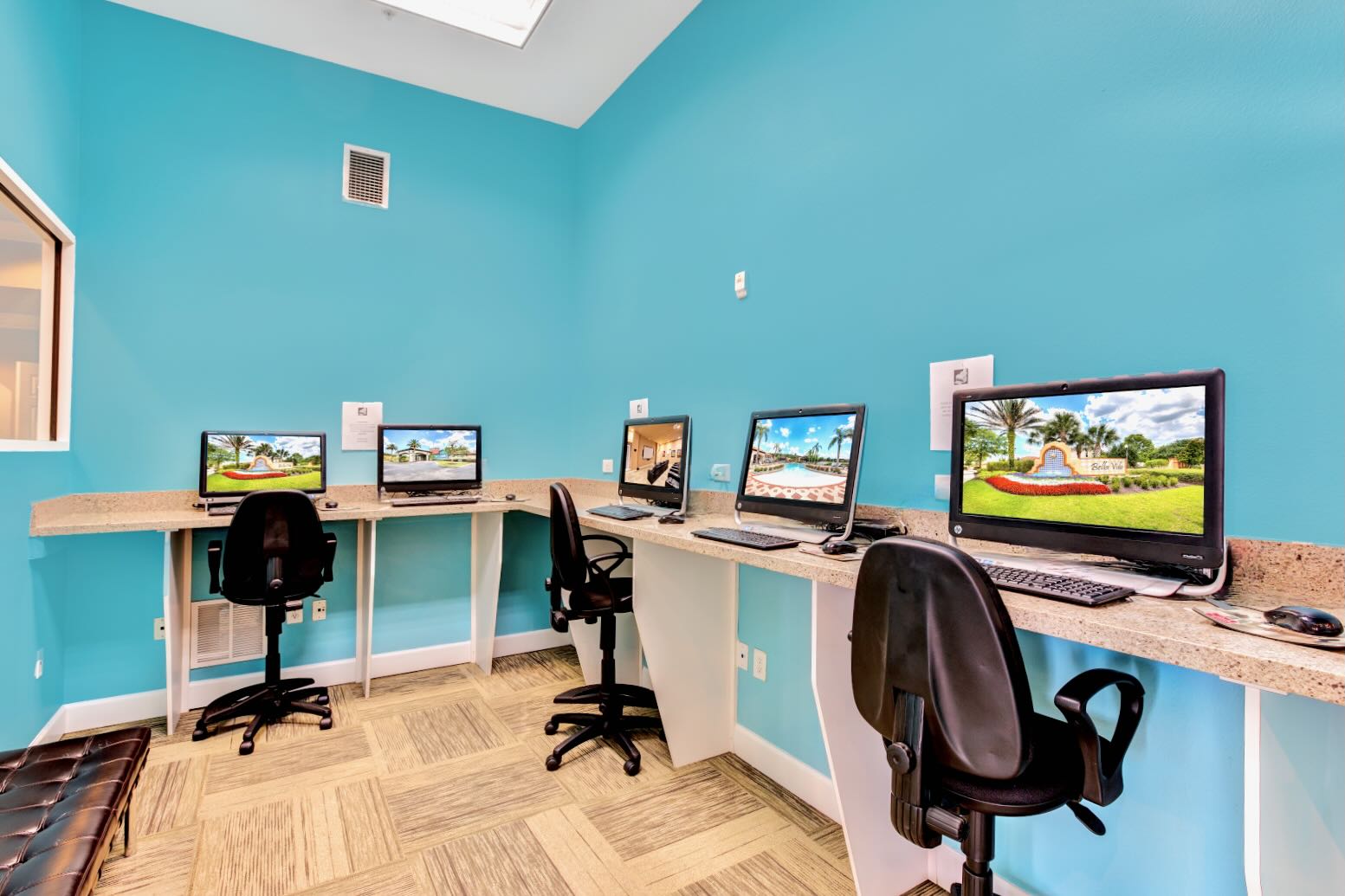 A modern communal computer room features bright blue walls and light patterned carpet, with multiple workstations equipped with desktop computers and ergonomic chairs. Long light-colored countertops provide ample workspace for guests.