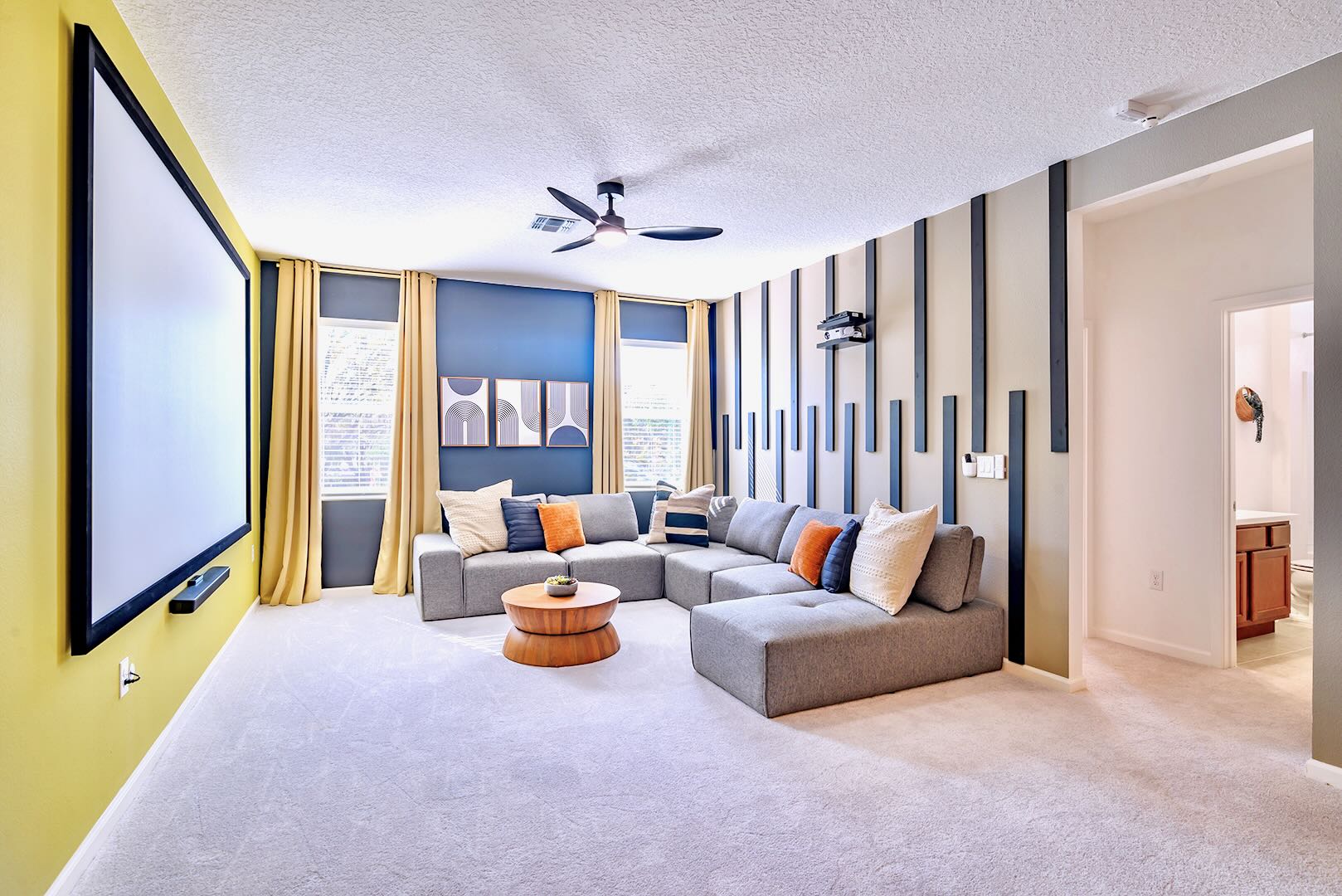 Modern media room with a large projection screen on a yellow accent wall, a spacious grey sectional sofa with colorful throw pillows, and a unique slatted accent wall with a projector.