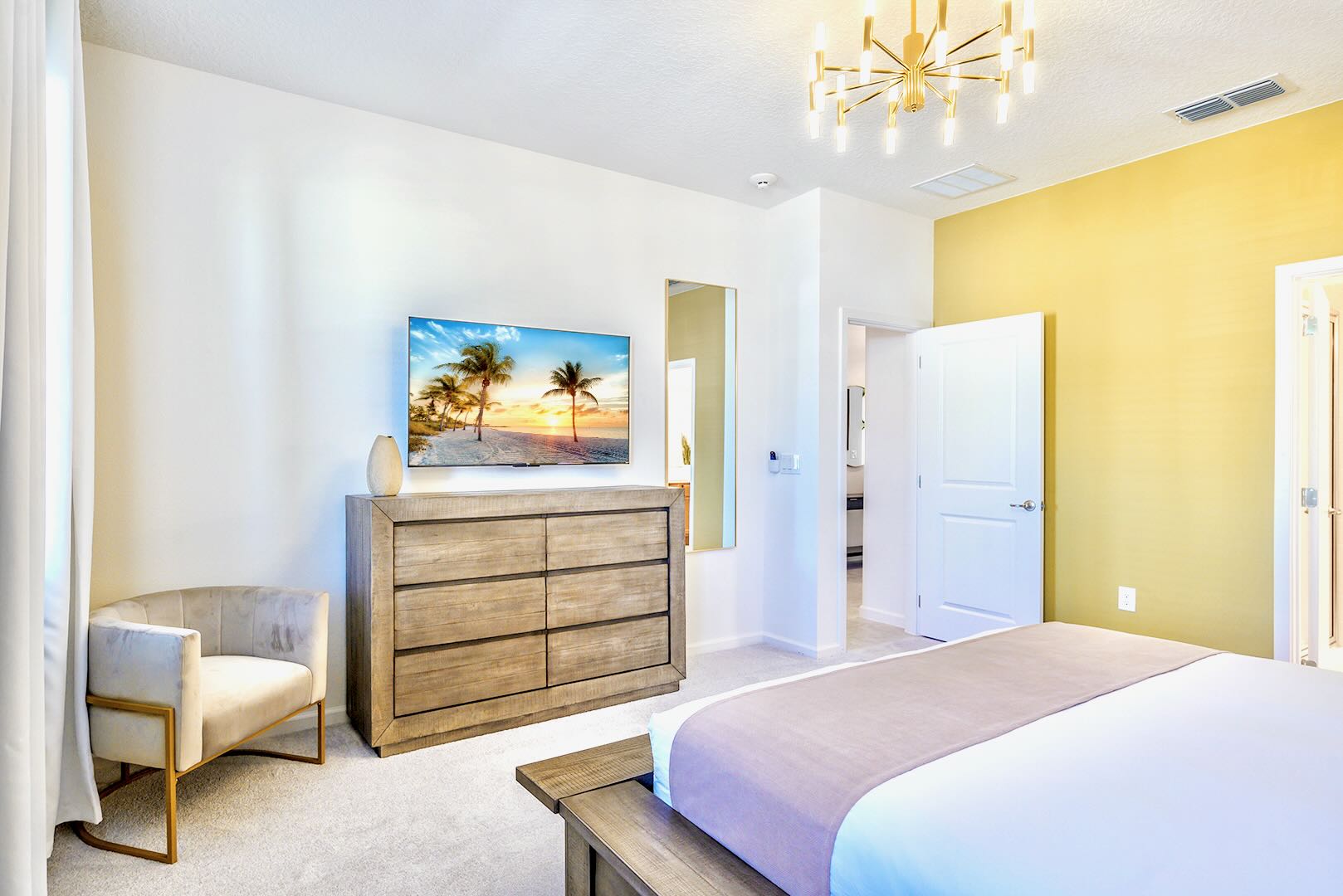 Bright bedroom featuring a large wooden dresser with a flat-screen TV displaying a beach scene, a plush armchair, and a full-length mirror, continuing the white and mustard yellow color scheme.