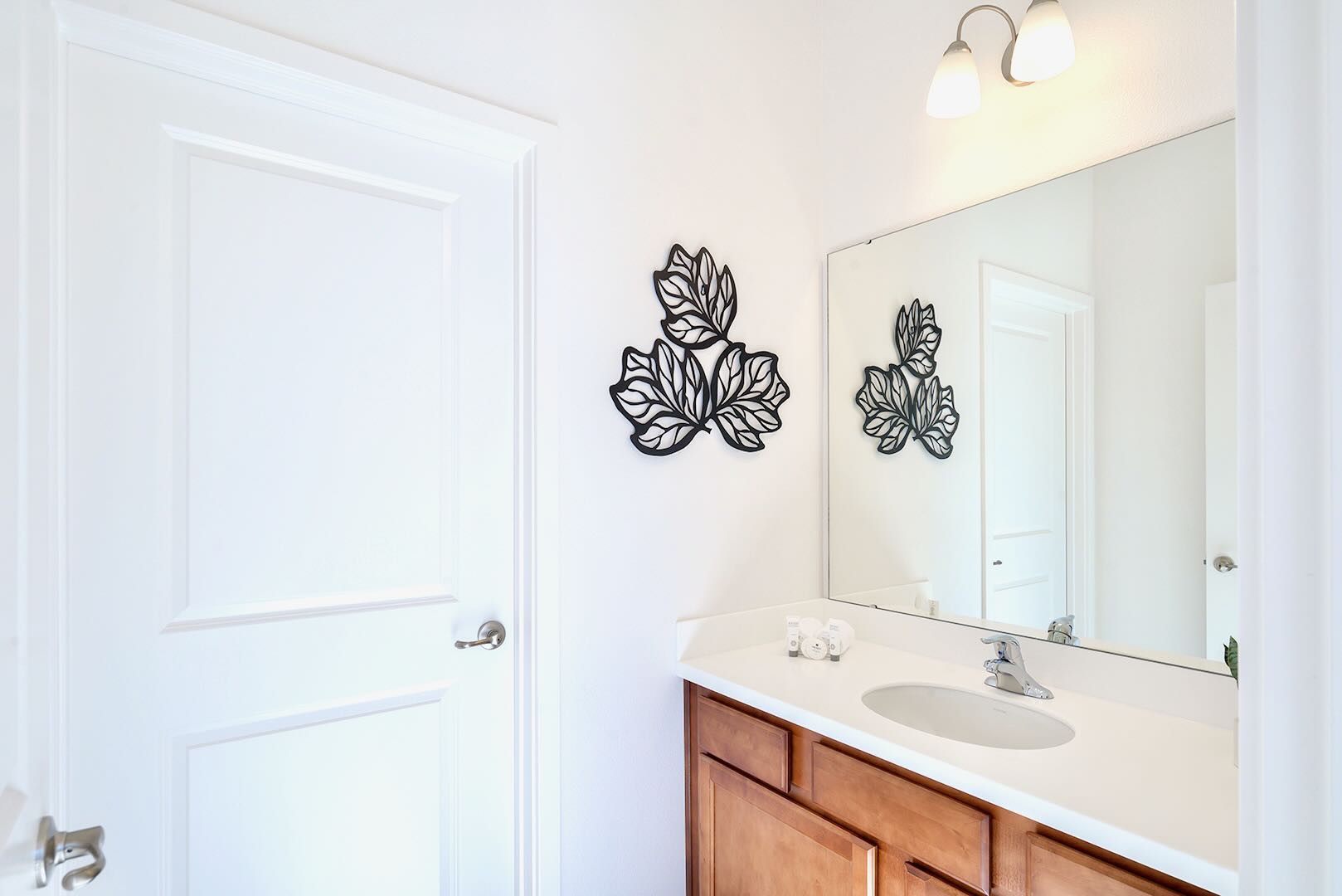 A bright bathroom featuring a wooden vanity with a white countertop and single sink, beneath a large mirror reflecting decorative metal leaf wall art. The room has clean white walls and a white paneled door.