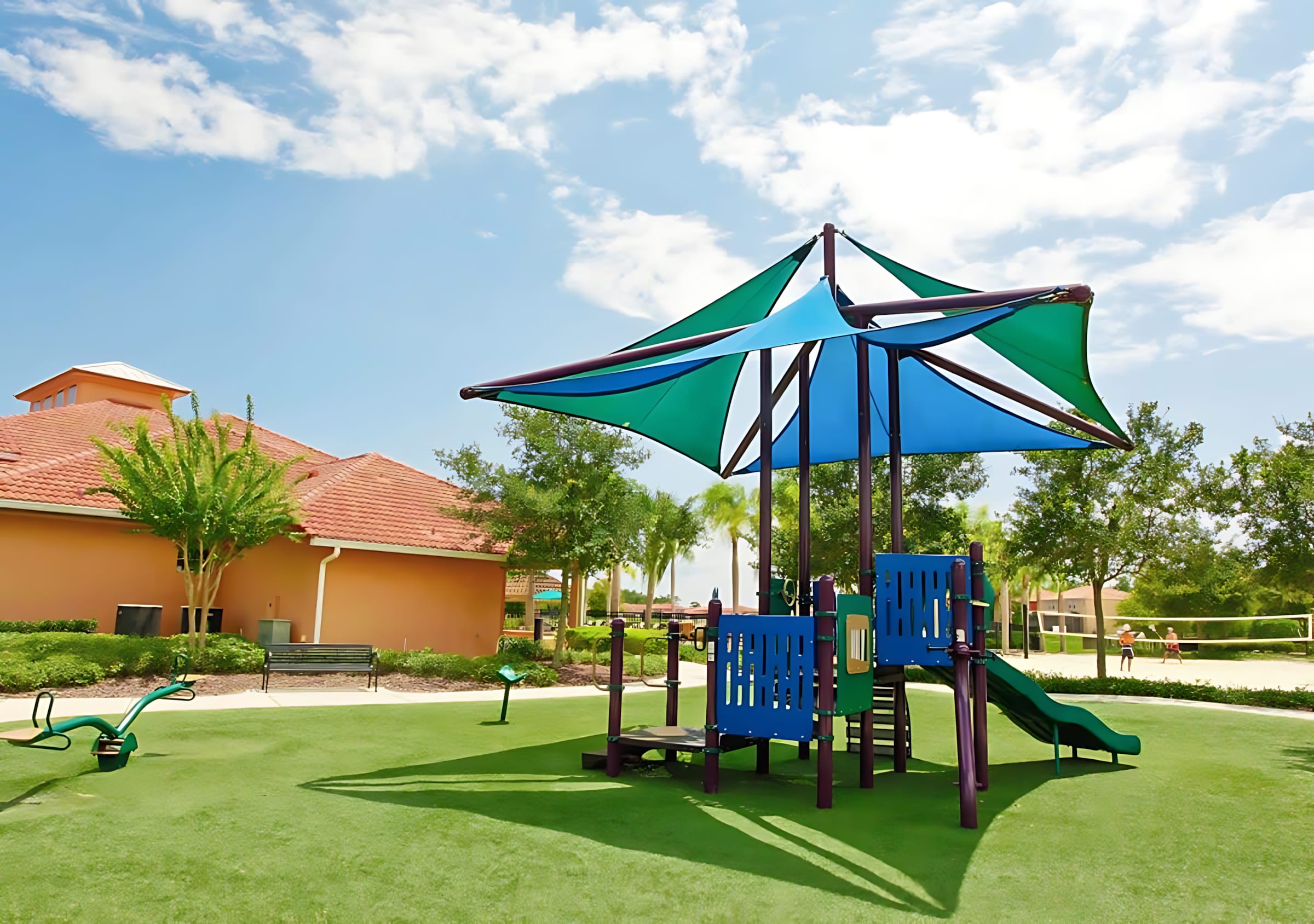 A vibrant outdoor playground features a blue and green shaded structure with slides and a seesaw, set on bright green artificial turf. A terracotta-roofed building and palm trees are visible in the background under a partly cloudy sky.