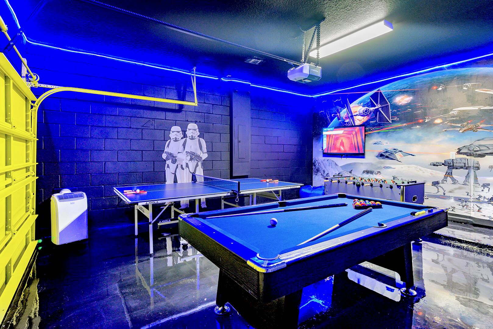 Dark-themed game room with vibrant blue LED strip lighting, featuring a blue felt pool table, a ping pong table, and a foosball table, all set against a Star Wars mural and black brick walls.