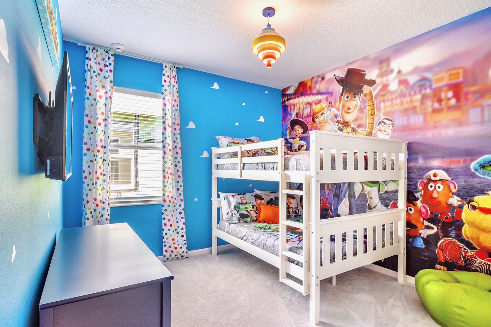 A colorful Toy Story themed bedroom with white bunk beds and a vibrant mural covering one wall depicting characters from the movie. The other walls are bright blue with cloud decals, and a dresser sits opposite the beds.