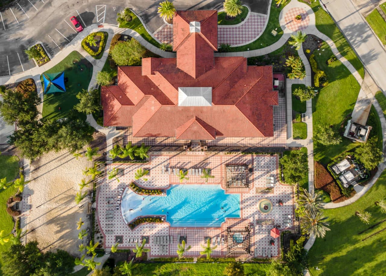 An overhead aerial view of a community clubhouse with a distinctive red tile roof, adjacent to a large swimming pool and surrounding amenities. The area includes lounge seating, pergolas, and green landscaping.