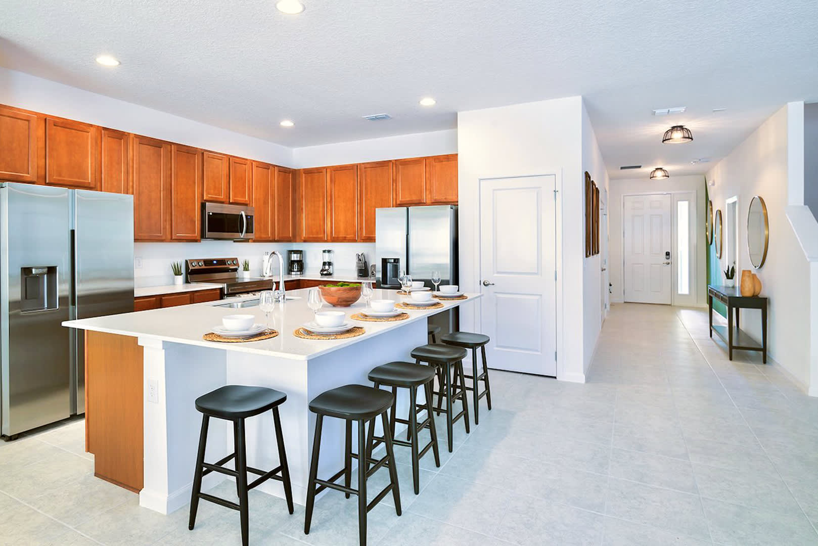Bright open-plan kitchen with light wood cabinets, white countertops, and stainless steel appliances, flowing into a tiled foyer with a console table and decorative mirrors, creating a welcoming entry.