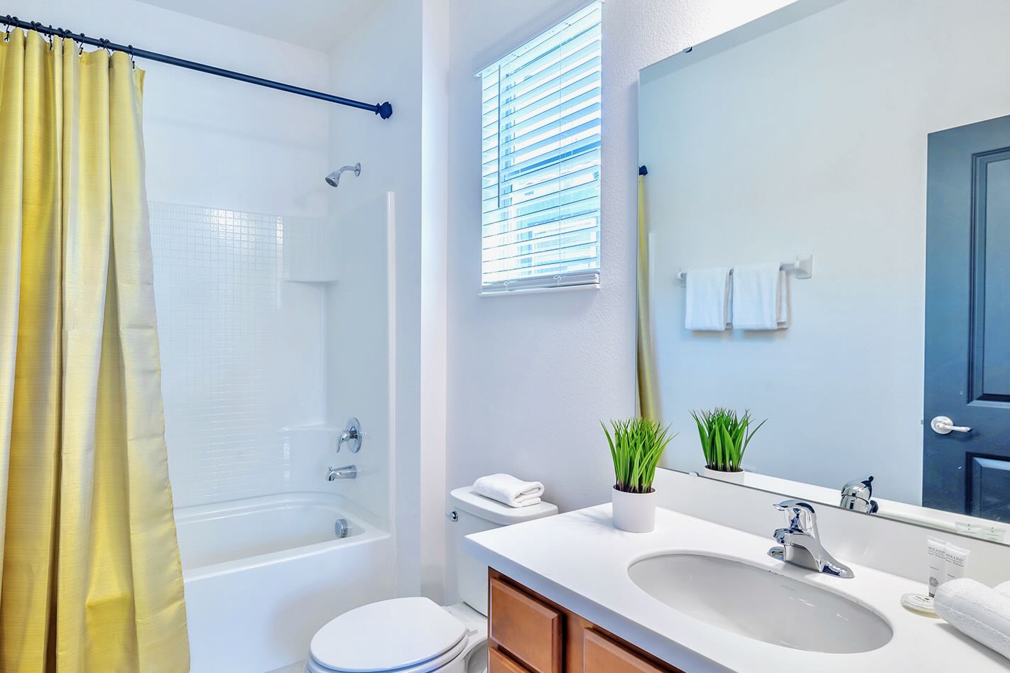 Bright bathroom featuring a white bathtub and shower combo with white tiled walls, a vibrant yellow shower curtain, and a single vanity with a white countertop and a small potted plant.