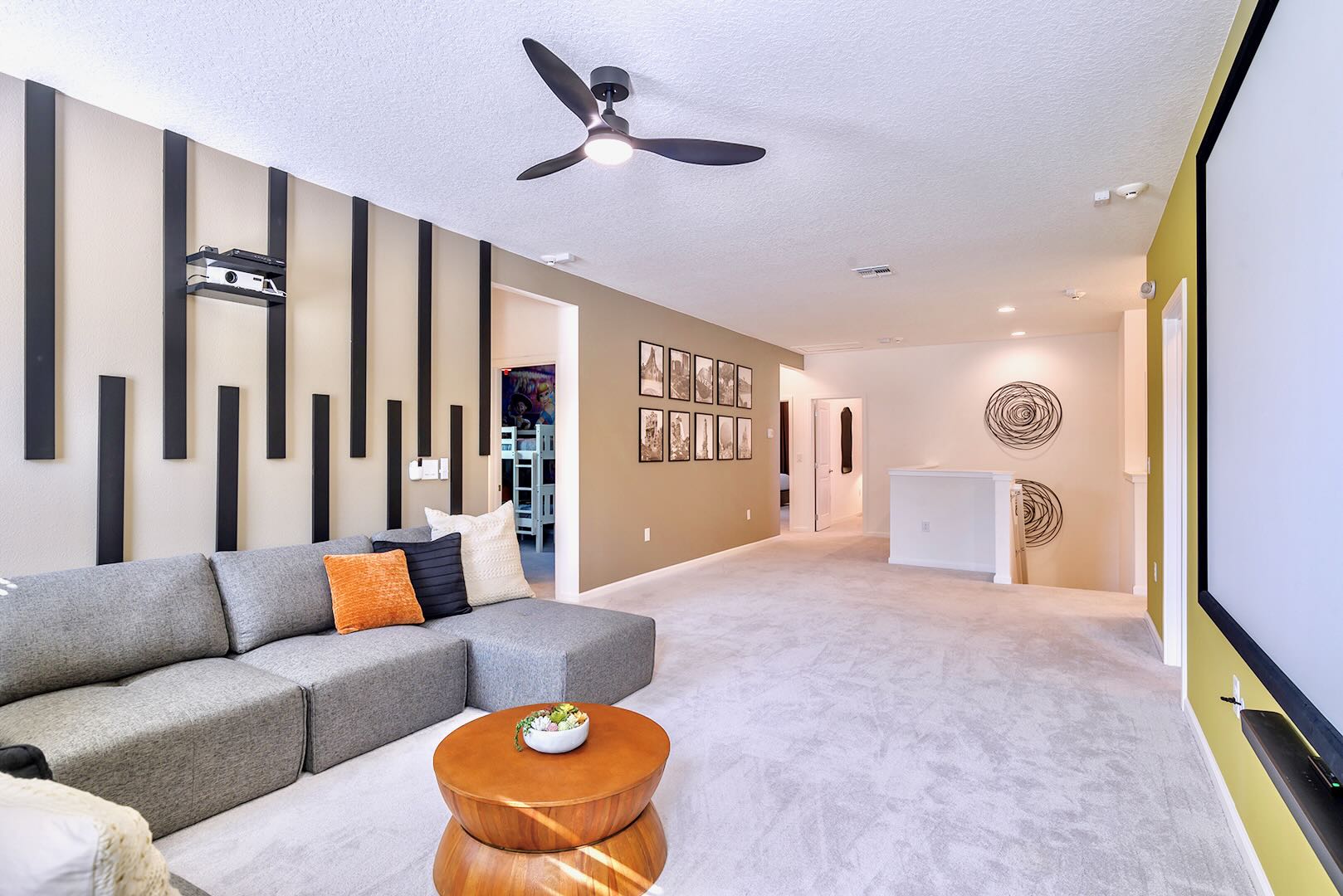 Contemporary media room featuring a grey sectional sofa, a round wooden coffee table, and a striking accent wall with vertical black slats, leading to a hallway with framed art and a staircase.