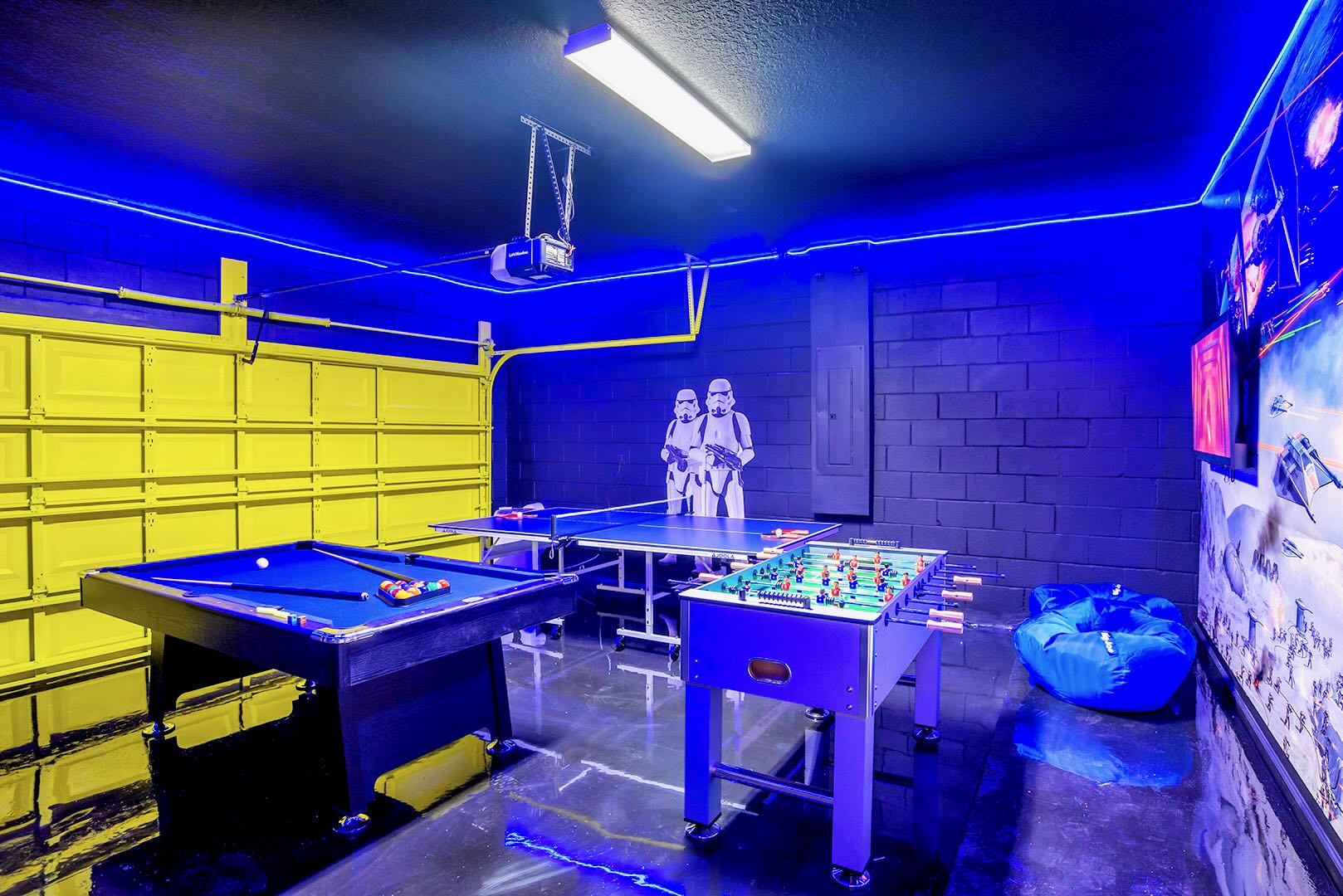 Star Wars-themed game room with bright blue LED lighting, featuring a blue pool table, a ping pong table, and a foosball table, with Stormtrooper cutouts and a yellow garage door.
