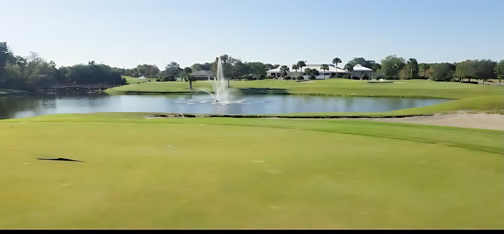 A scenic view of a sprawling green golf course features a large pond with a decorative fountain spraying water high into the air. In the distance, residential buildings and lush trees frame the horizon under a clear, sunny sky.