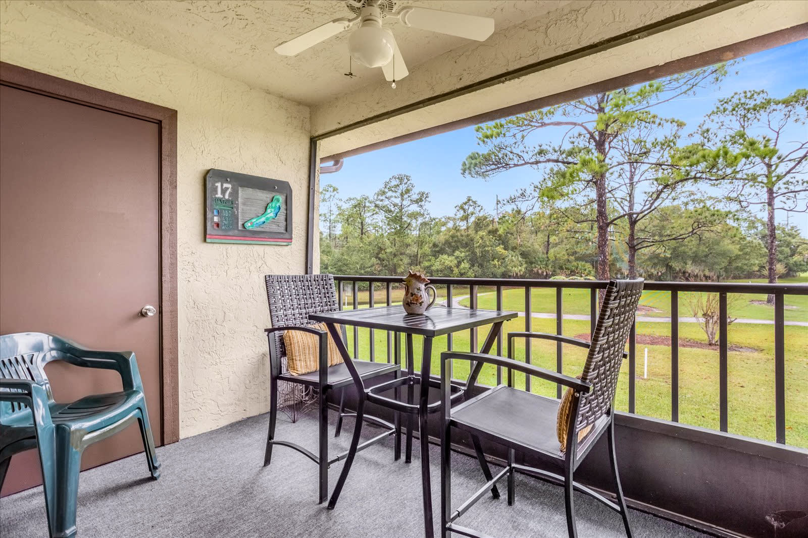 A screened balcony with a ceiling fan and a small black metal table with two chairs, offering a view of a golf course and pine trees, with a golf hole marker on the wall.