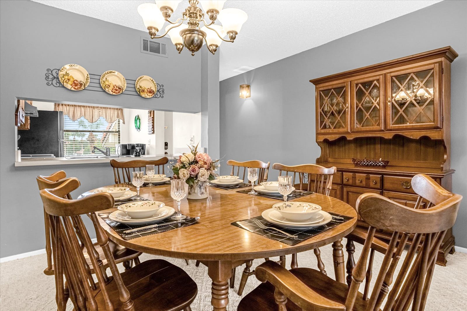 A dining area with a round wooden table set for a meal, surrounded by wooden spindle-back chairs, adjacent to a pass-through window to the kitchen, and a wooden hutch against a gray wall.