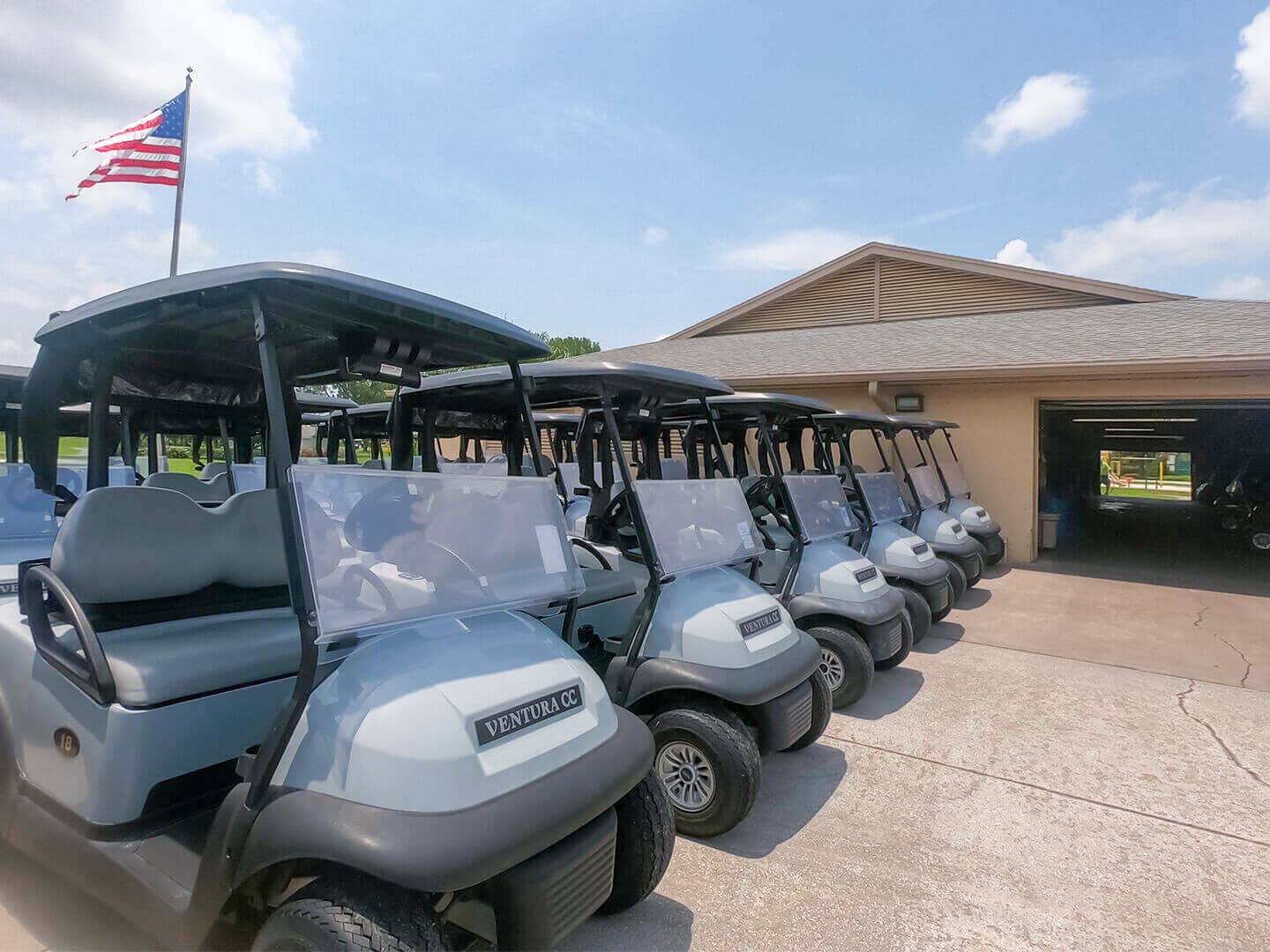 A line of light gray golf carts, each branded 'VENTURA CC,' are neatly parked on a paved surface outside a building, likely a clubhouse. An American flag waves proudly in the background under a bright blue sky.