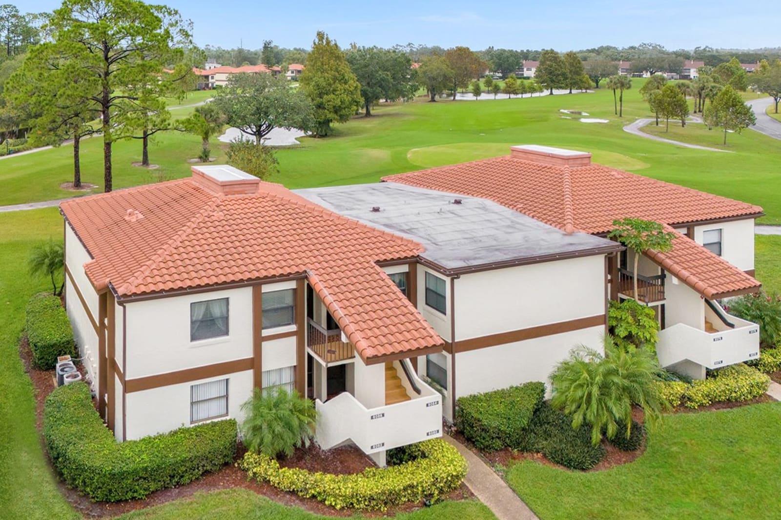 An aerial view showcases a multi-story residential building with light-colored stucco walls and distinctive terracotta-style roof tiles. The property is surrounded by manicured green landscaping and borders a sprawling golf course under a clear sky.
