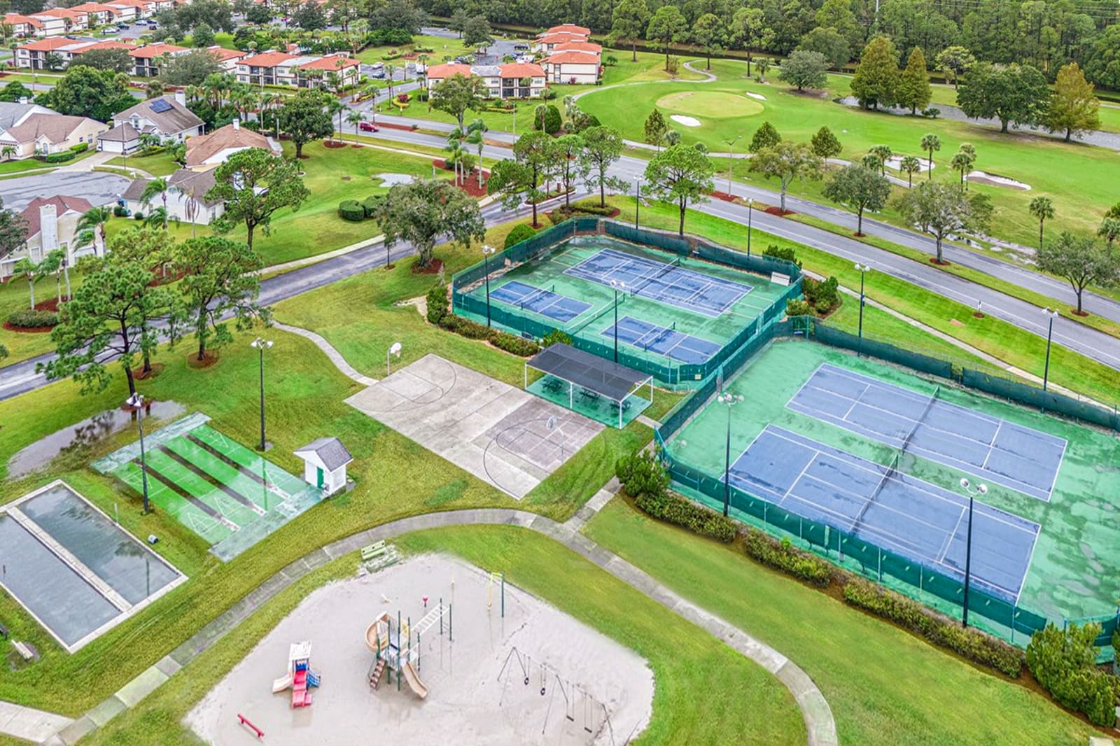 An aerial perspective reveals extensive outdoor recreational facilities, including multiple blue tennis courts, a concrete basketball court, bocce ball courts, and a children's playground. These amenities are surrounded by manicured lawns and trees, with residential buildings in the background.