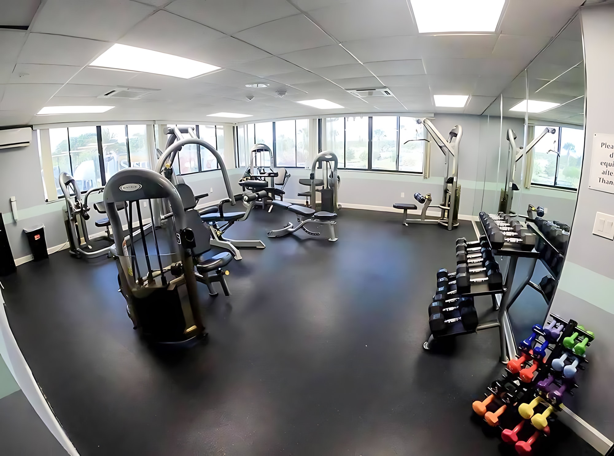 A well-equipped communal gym featuring various weight machines, free weights on a rack, and a large mirror wall. The room has a dark rubber floor, bright overhead lighting, and large windows offering natural light.