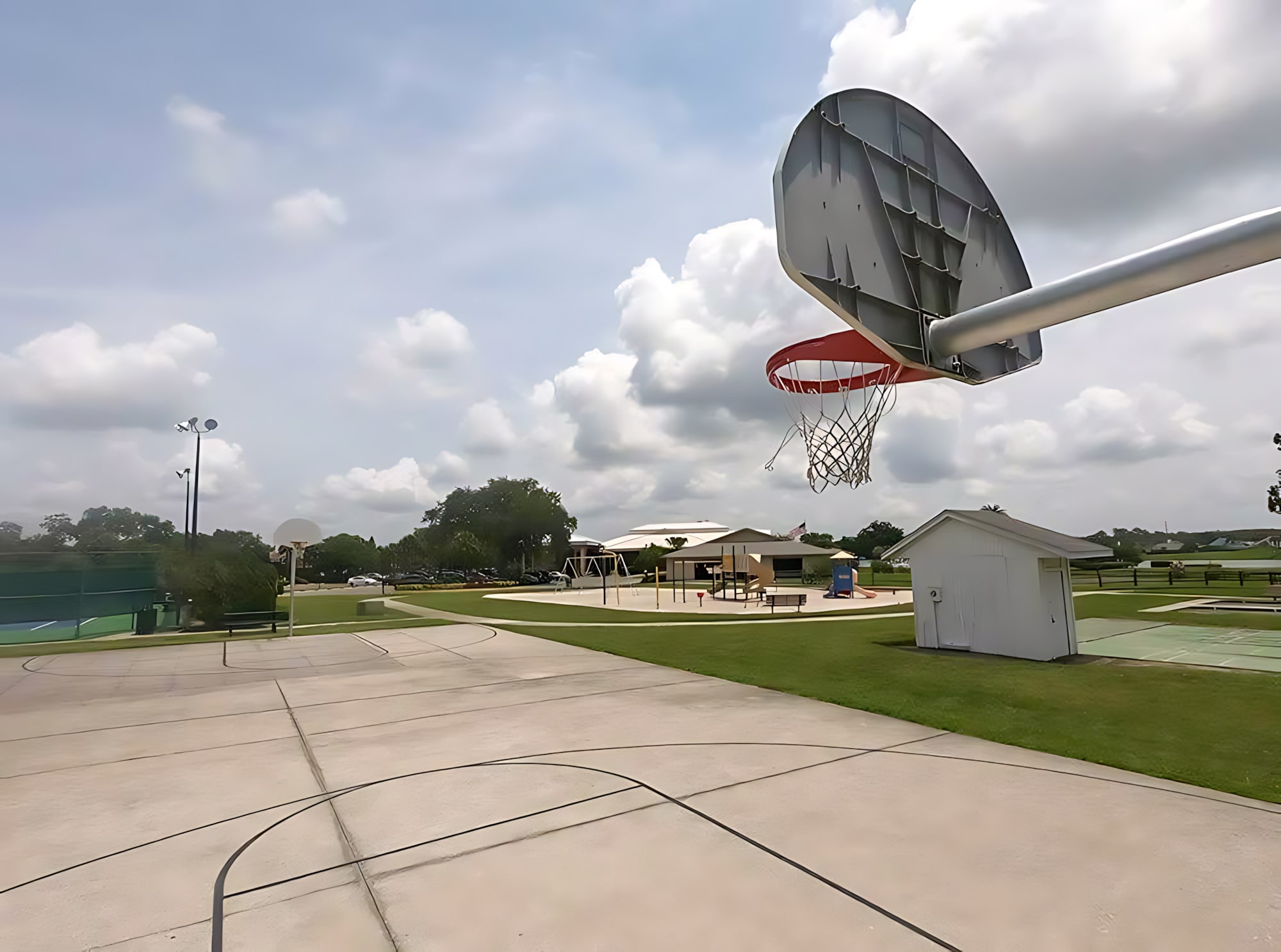 An outdoor concrete basketball court with a hoop in the foreground is visible under a partly cloudy sky. In the background, other recreational amenities like a playground and possibly tennis courts are nestled within green spaces.