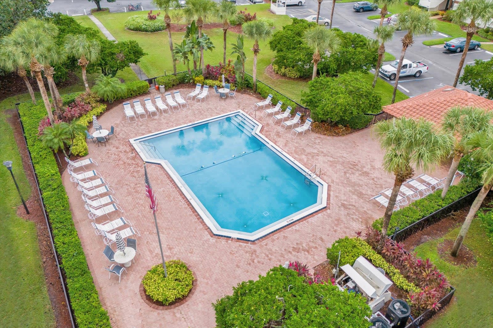An aerial shot captures a large, geometrically shaped outdoor swimming pool surrounded by a spacious red brick patio. Numerous white lounge chairs and tables with umbrellas are arranged around the pool, with palm trees and lush foliage providing a tropical setting.